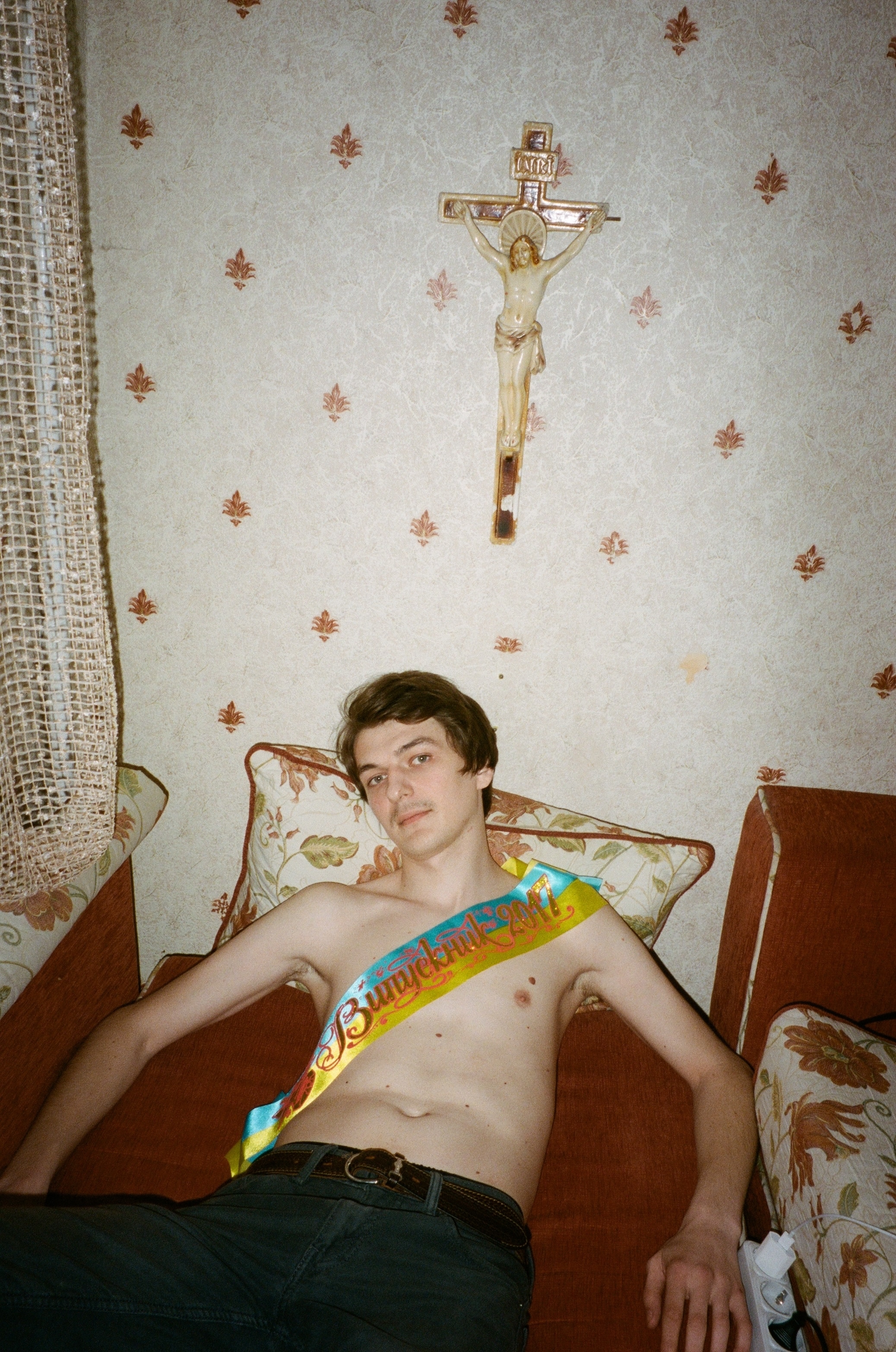 A young man lying on a floral-patterned sofa, holding a snack bar, with a crucifix on the wall above.
