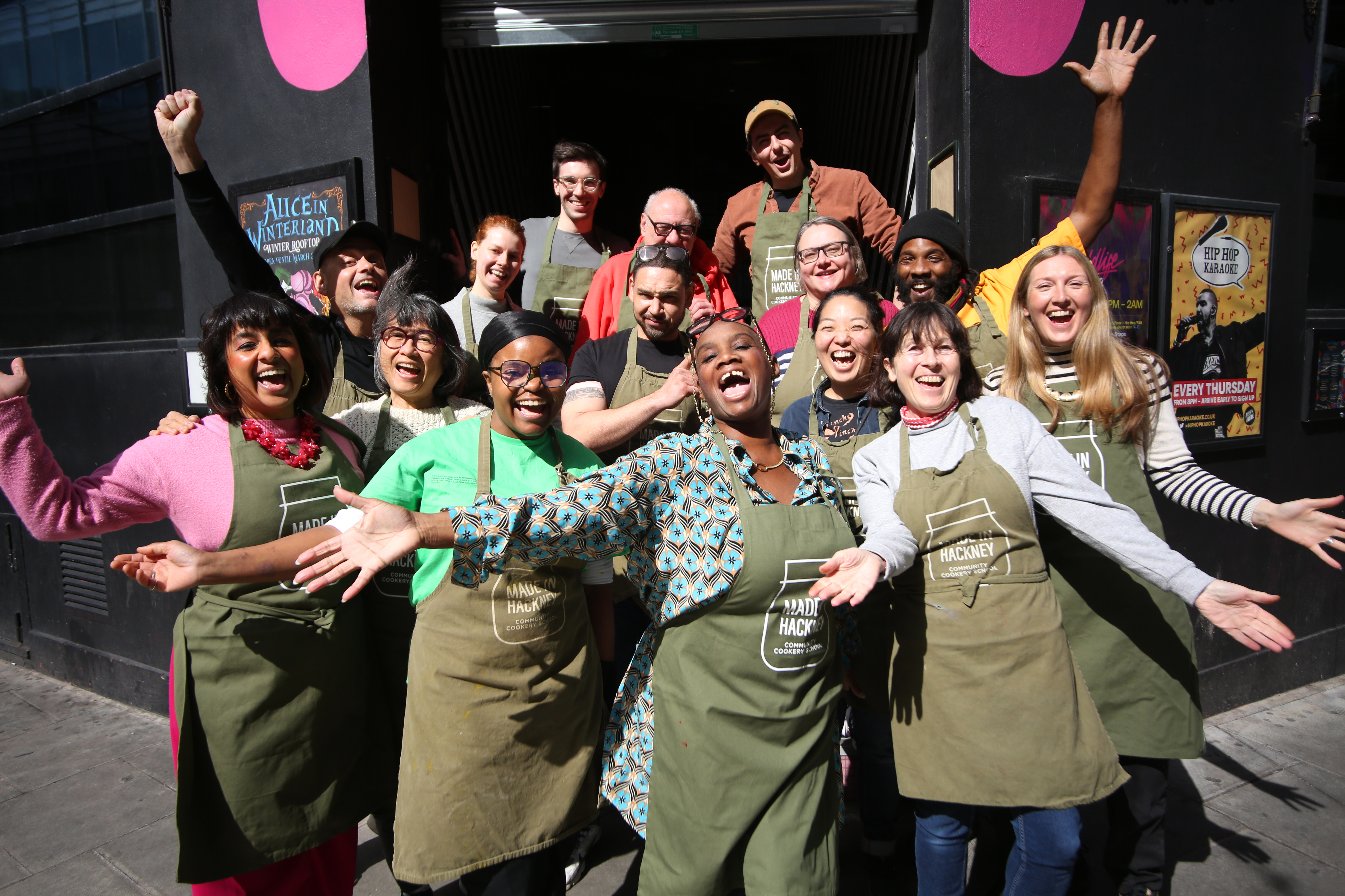 Large group of people wearing brown aprons celebrating with raised arms outside shopfront with pink balloons and signage.