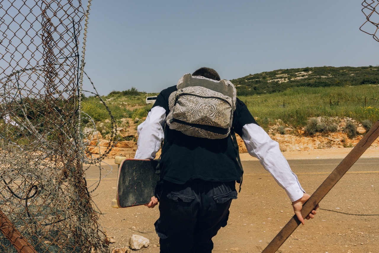 Backpack-wearing person with hiking attire walking along a dirt path surrounded by scrubland and wire fence.