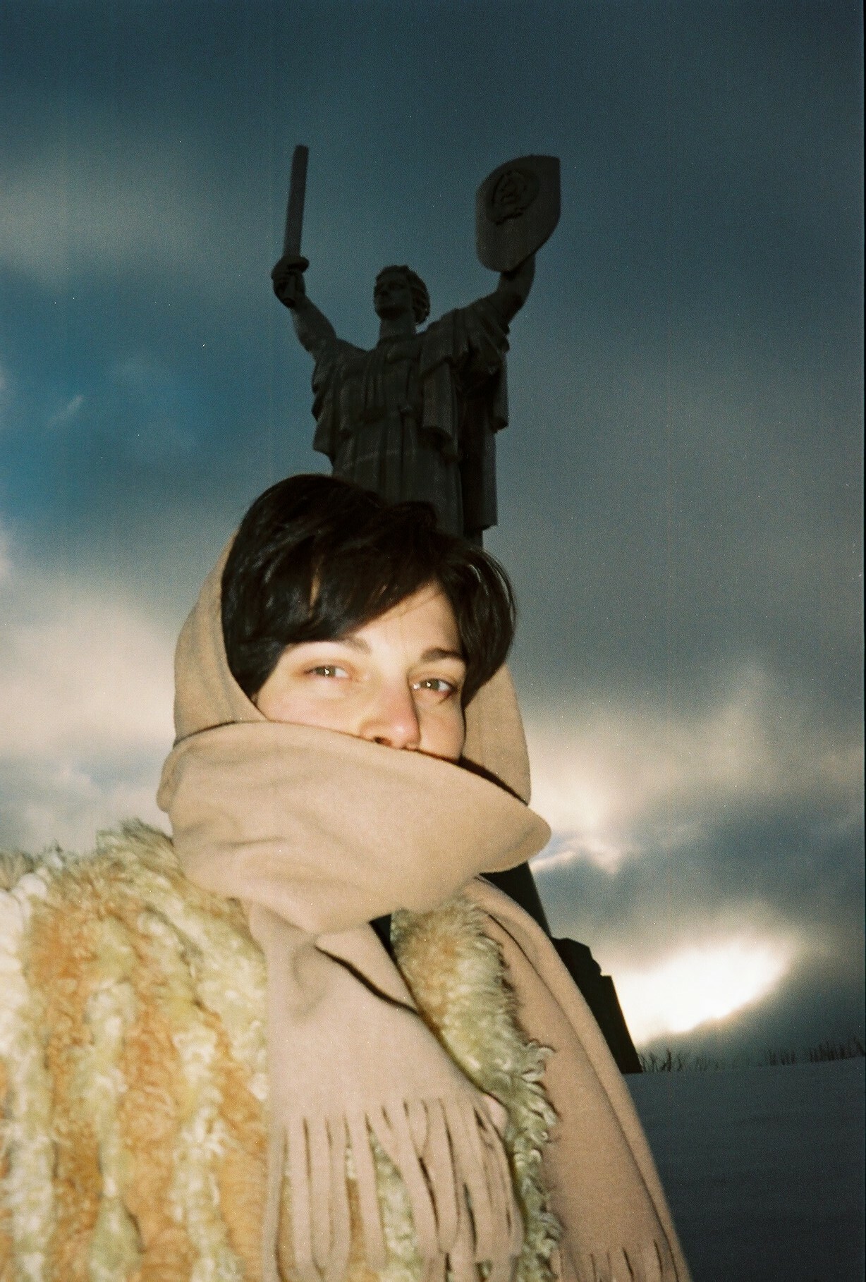 Person wrapped in a beige blanket holding a sword-wielding statue in the background.