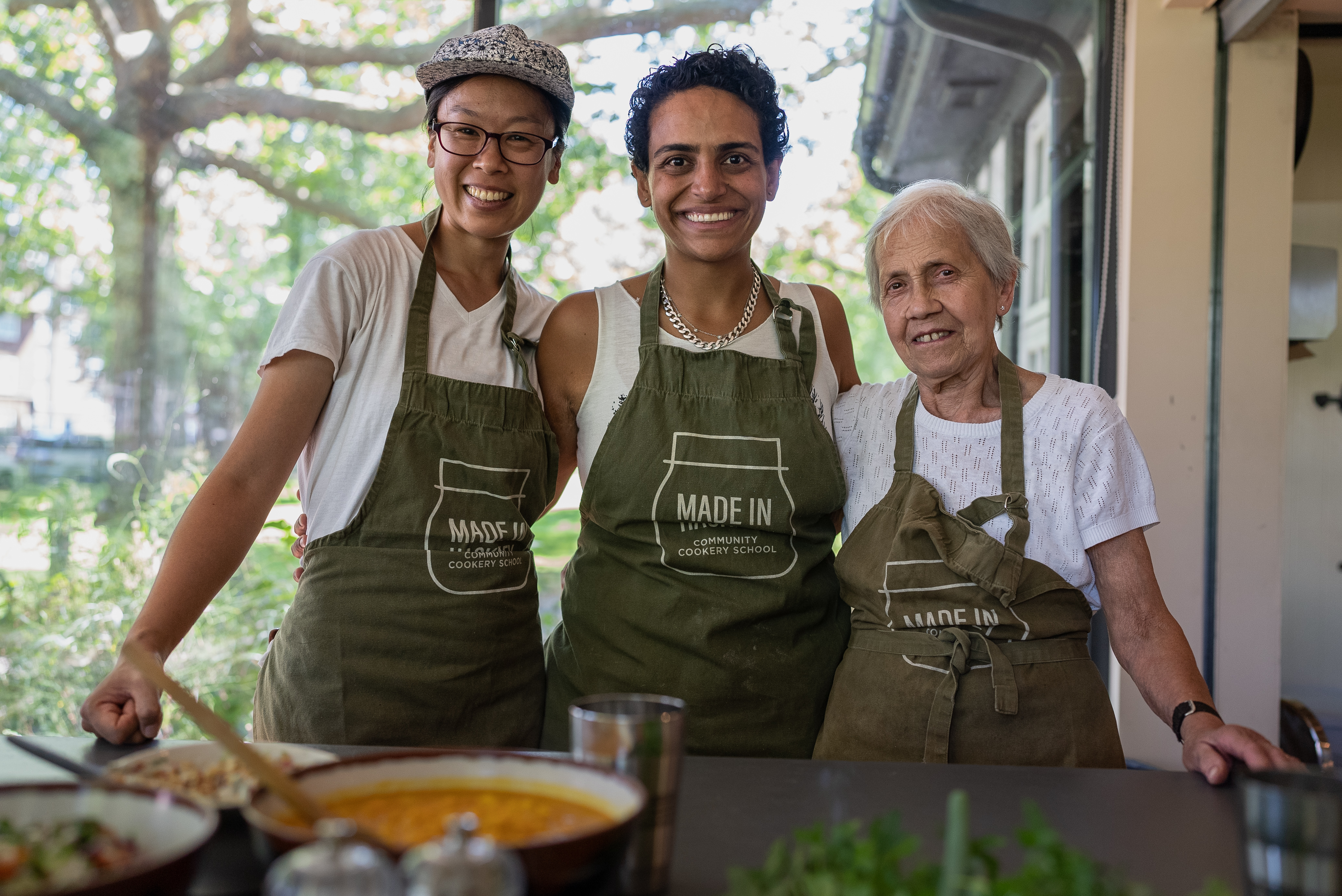 Three people wearing green aprons with "Made In" text standing together in food truck or mobile kitchen, smiling at camera.