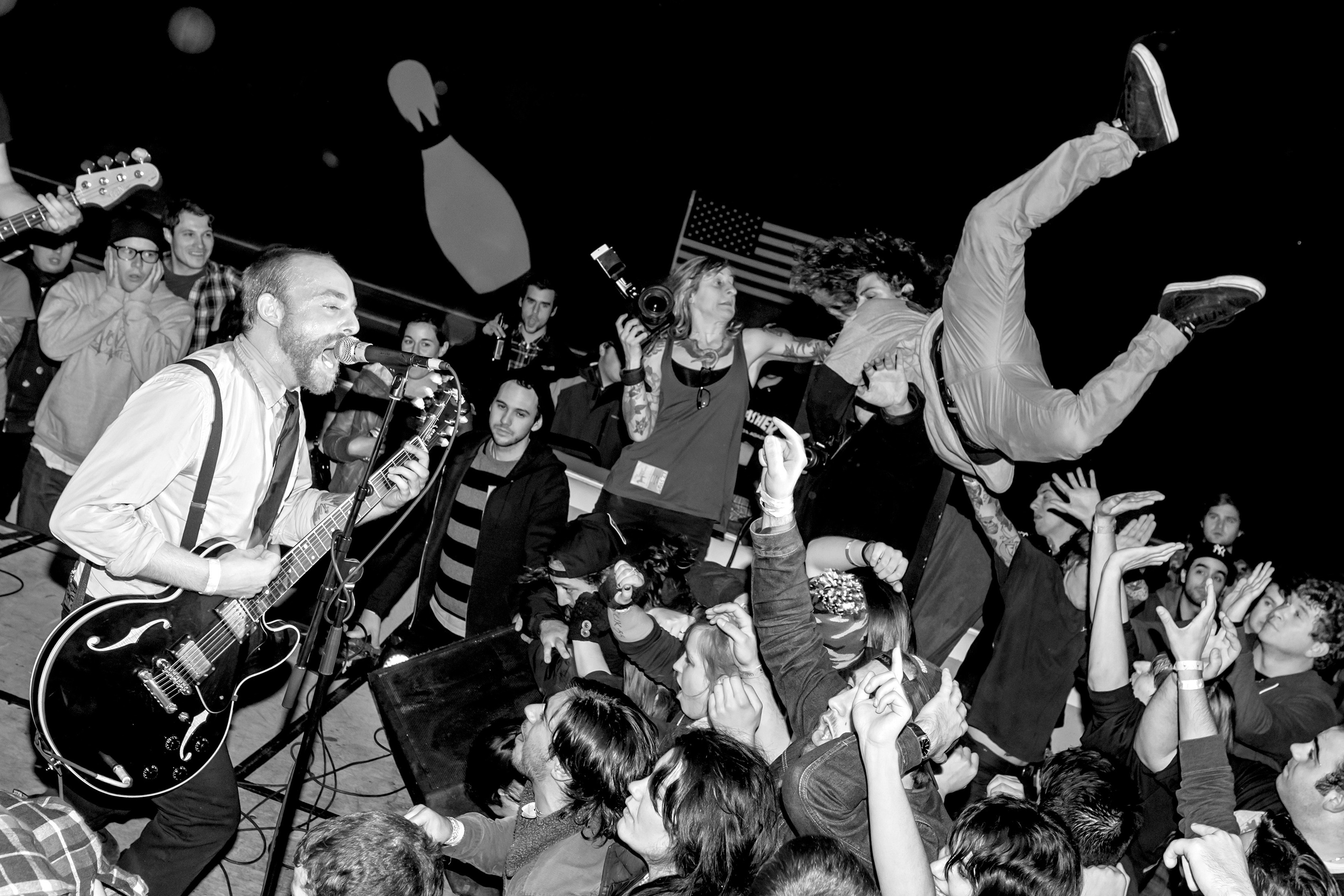 Black and white image of guitarist performing on stage with energetic crowd below, people with raised arms and American flag visible.