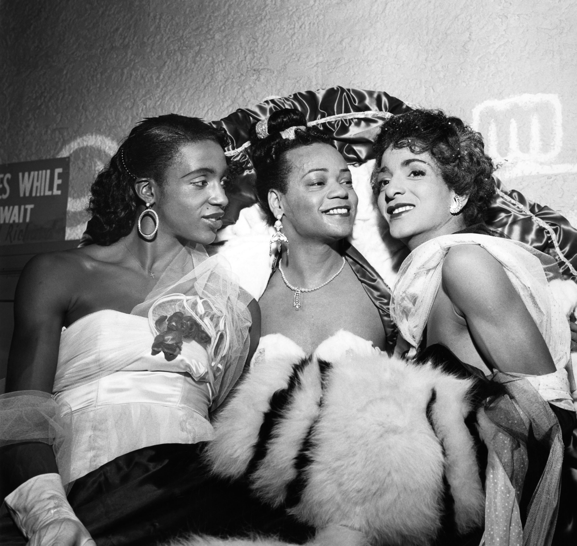 Three smiling women wearing glamorous dresses and fur coats posing together.
