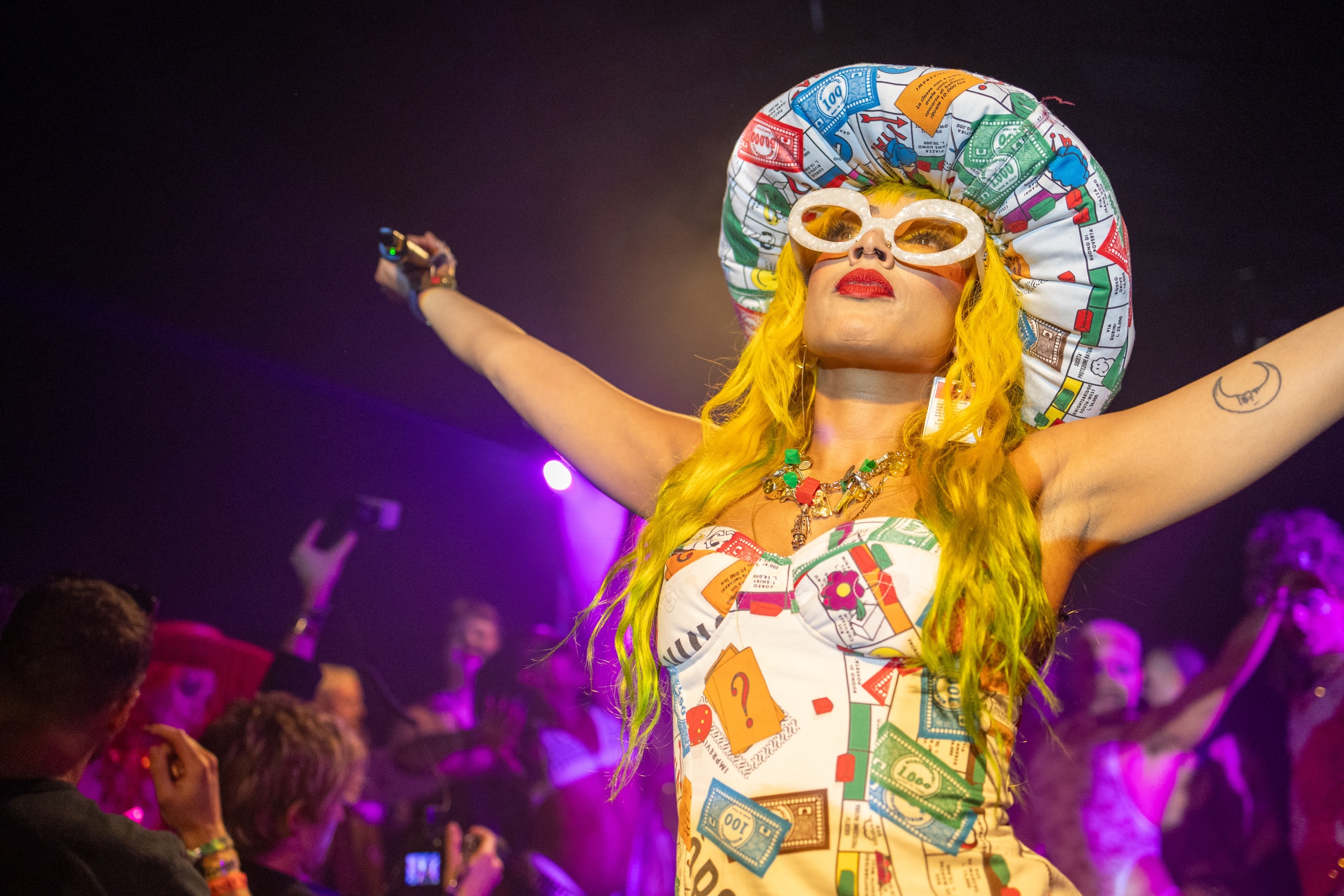 Performer on stage wearing colourful costume with large headdress and sunglasses, arms raised in expressive gesture, surrounded by cheering crowd.