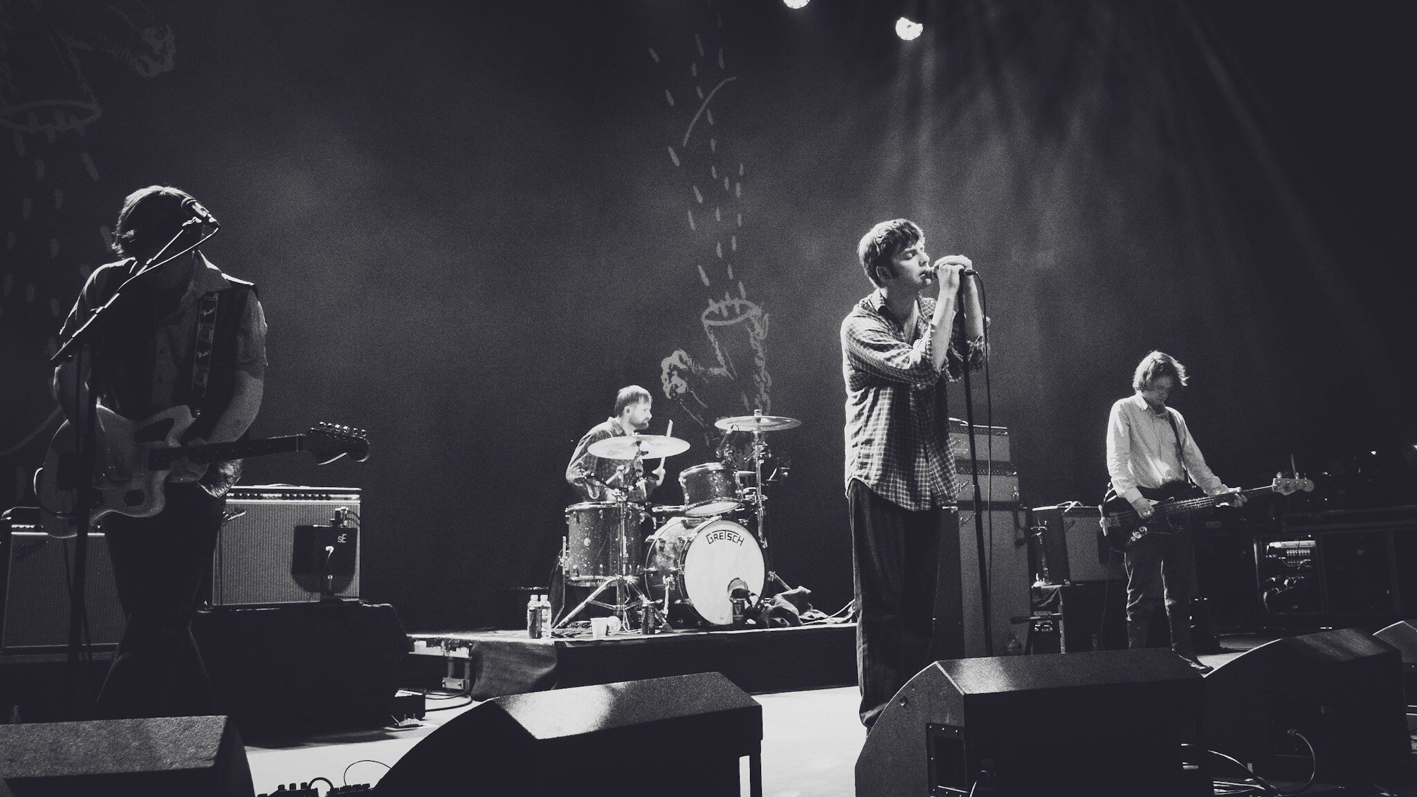 Four-piece band performing on dimly lit stage with dramatic spotlighting, singer at centre microphone, drummer, guitarist, and bassist visible.