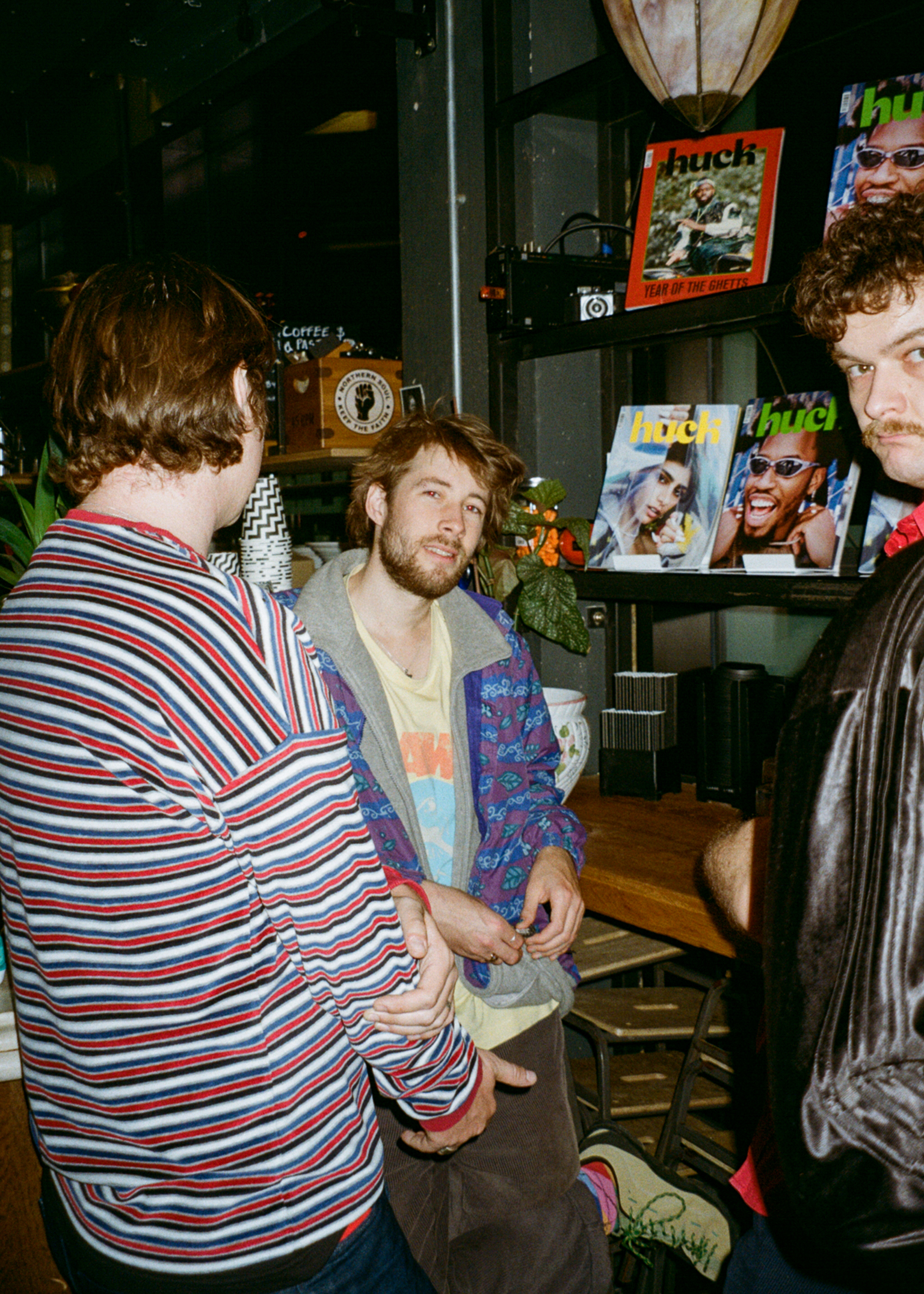 Three young men in casual clothing inside a record shop with magazine racks and shelving visible in background.