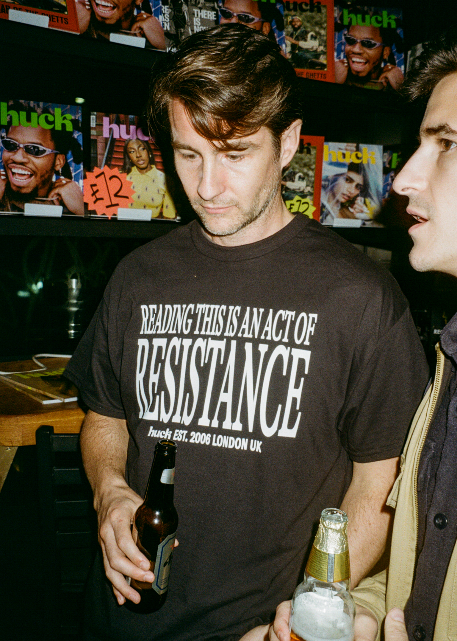 Man in dark t-shirt with "Reading this is an act of resistance" text, standing near magazines and posters on wall behind him.