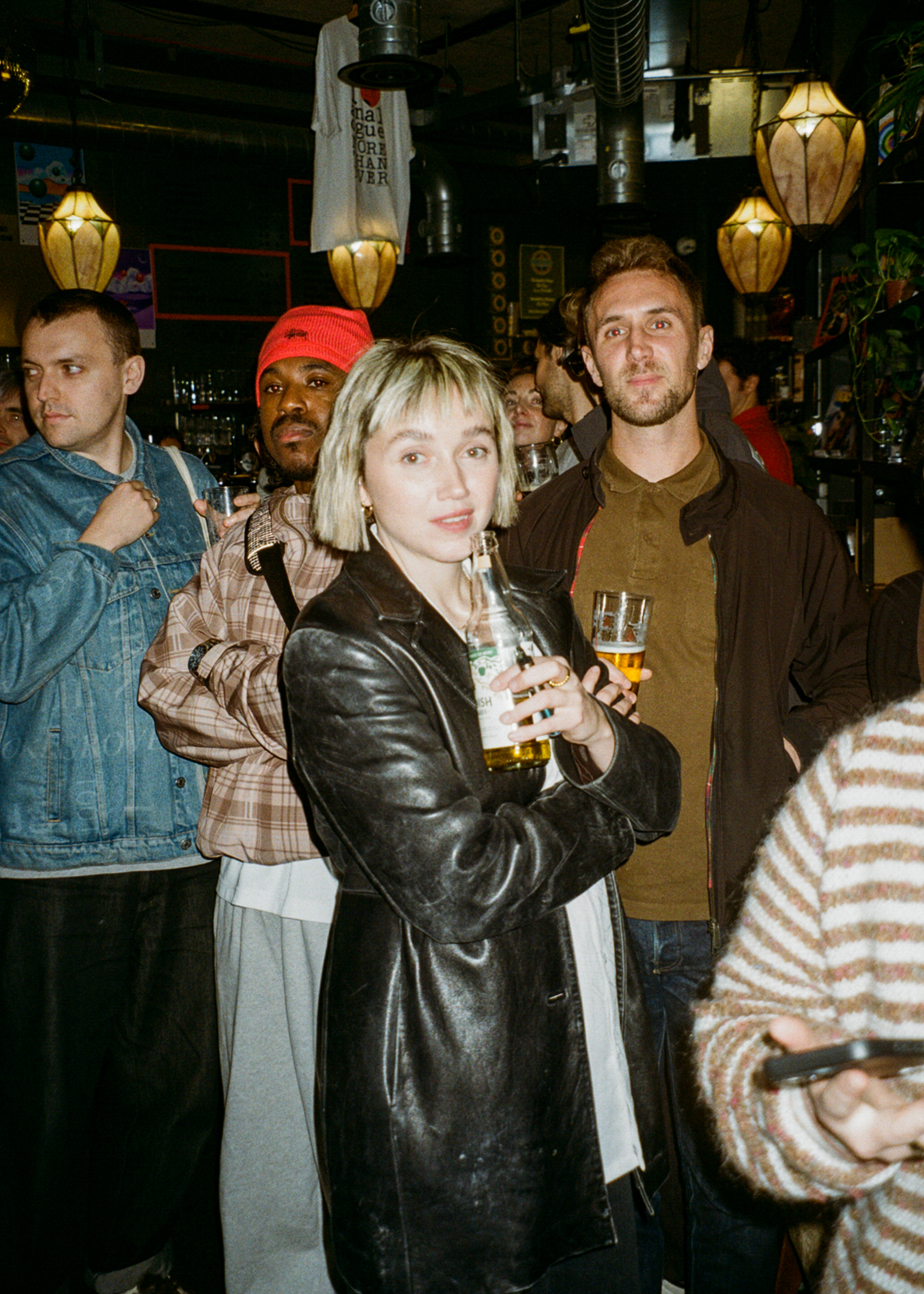 Group of people in dimly lit pub with hanging lanterns. Woman in black leather jacket holds beer glass, surrounded by men in casual clothing.