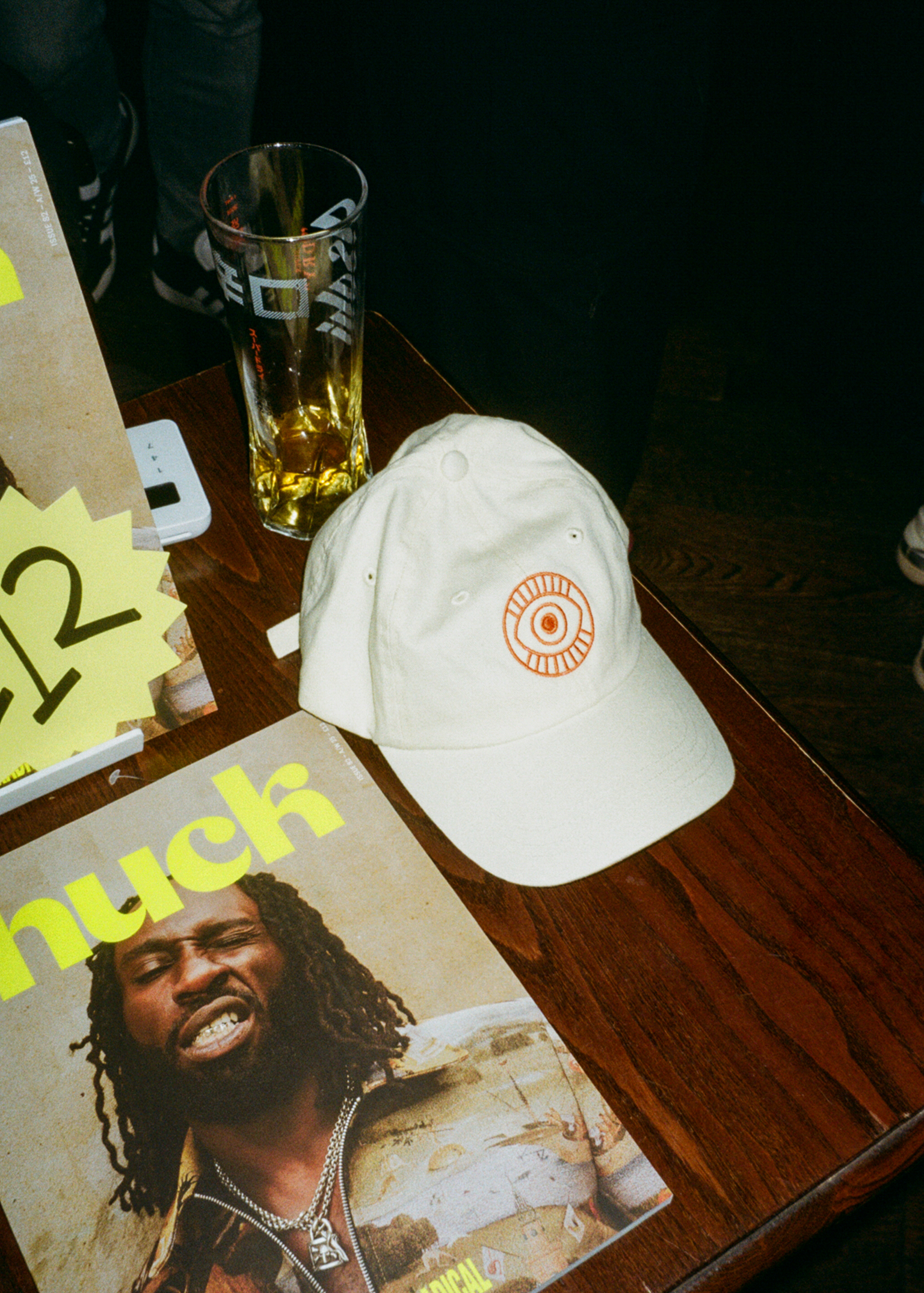 White baseball cap with orange circular logo, glass with amber liquid, magazine, and yellow sticky notes on wooden table surface.