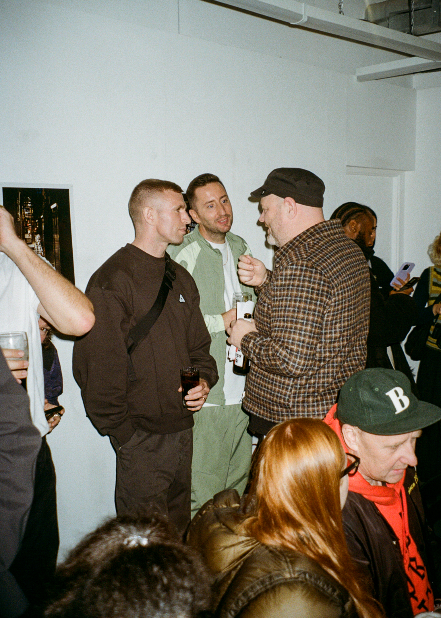 Group of people socialising at indoor gathering, man in brown jumper talking to man in green cap and checked shirt, white walls visible.
