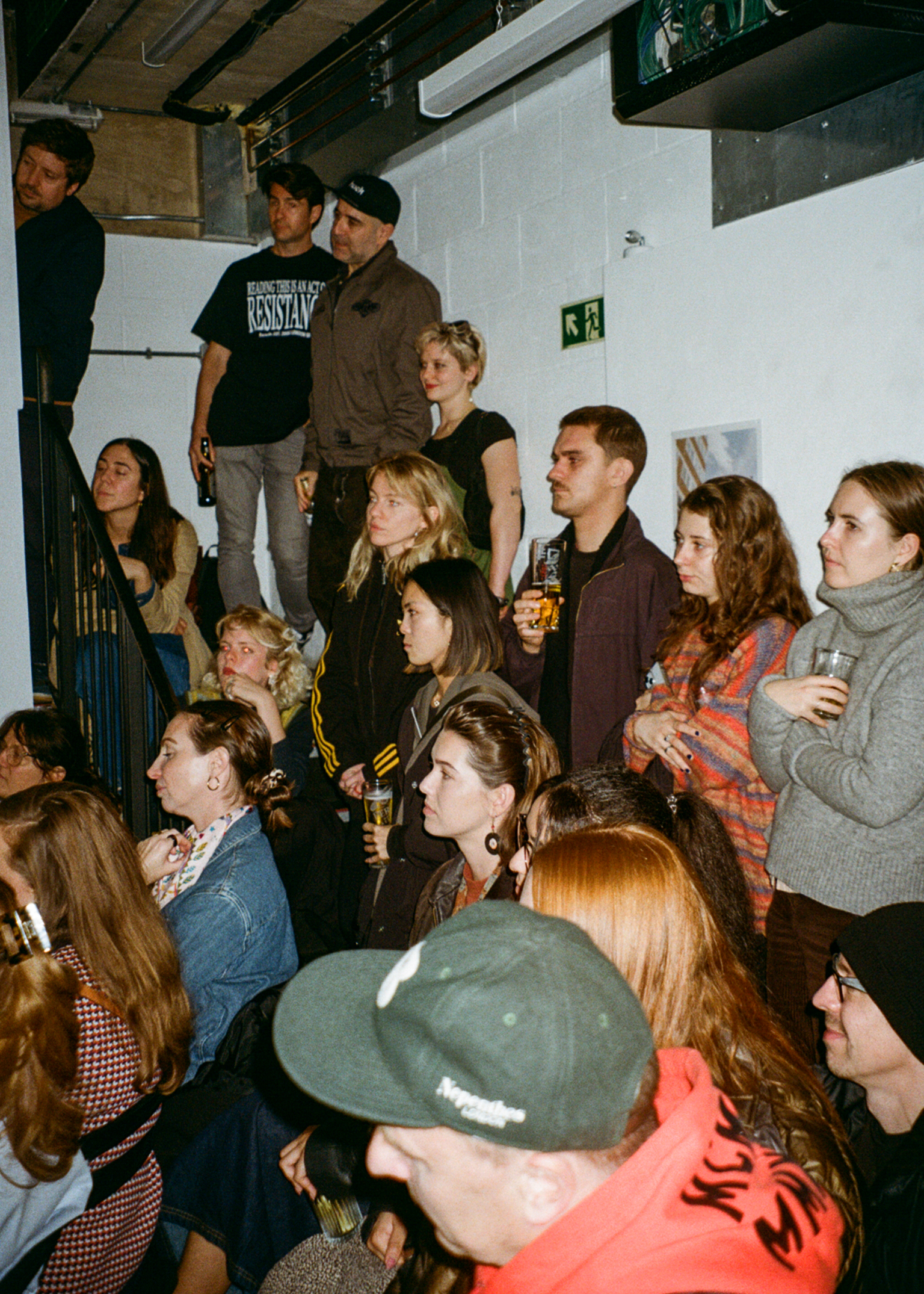 Group of young people gathered in cramped white-walled room with exposed ceiling beams, some sitting, others standing, holding drinks.