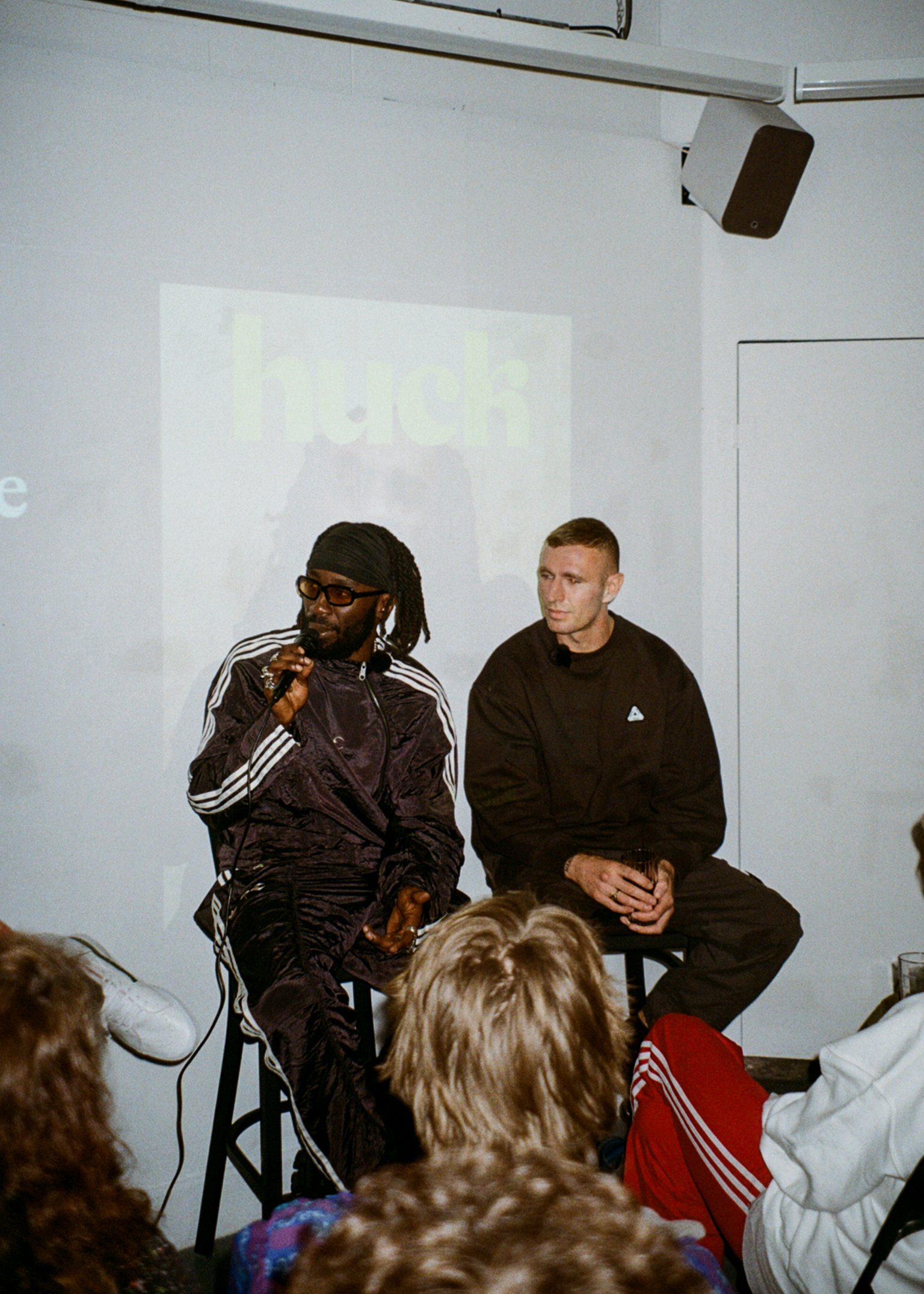 Two men seated on stools during presentation, one in dark tracksuit speaking into microphone, audience members visible in foreground.