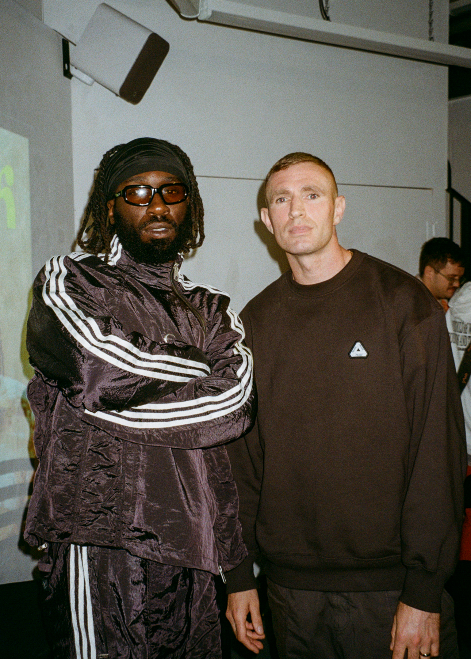 Two men standing together indoors - one wearing dark tracksuit with white stripes and sunglasses, other in brown jumper.