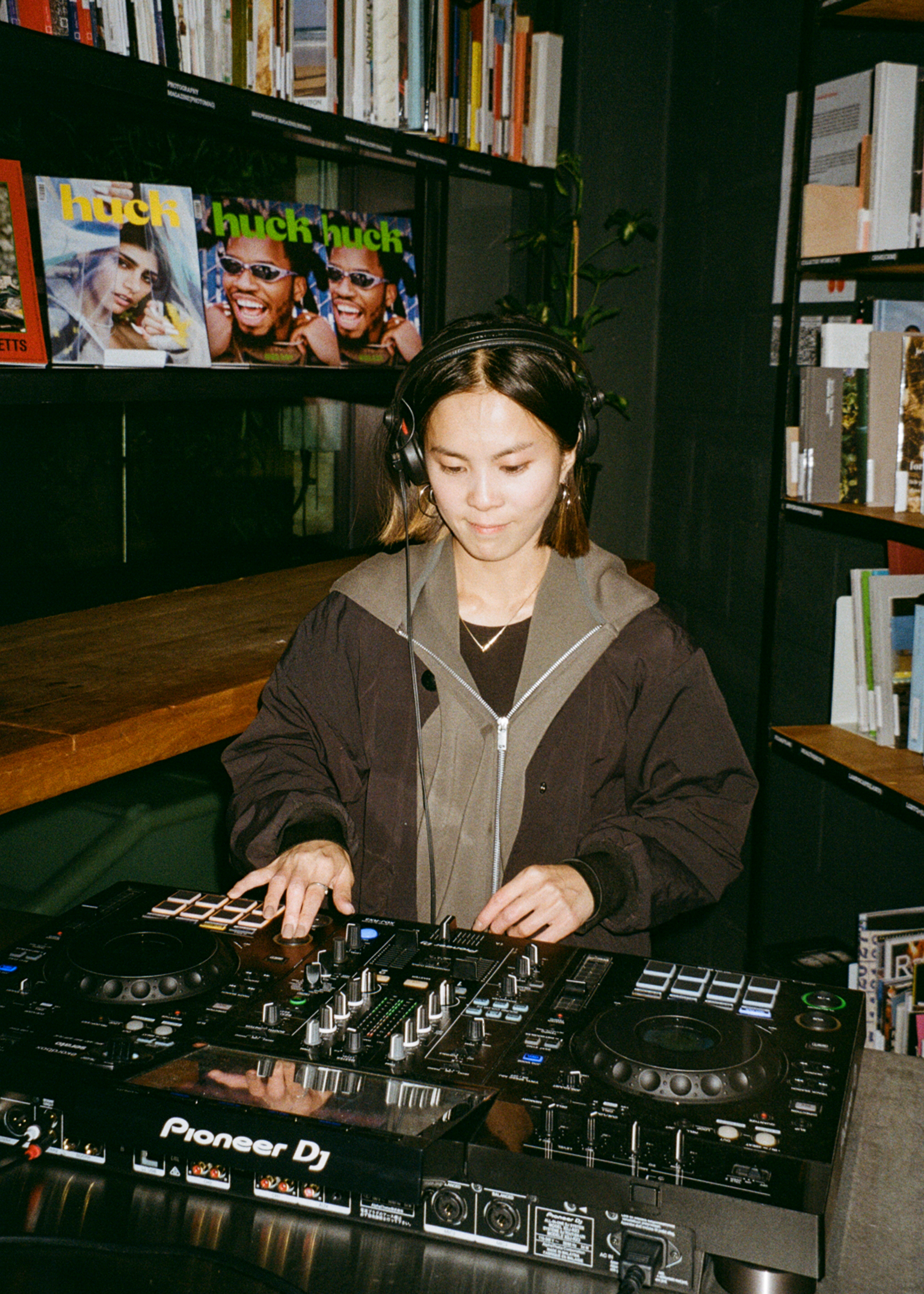 Woman wearing headphones and brown hoodie operating Pioneer DJ controller in bookshop with shelves and magazines visible behind her.