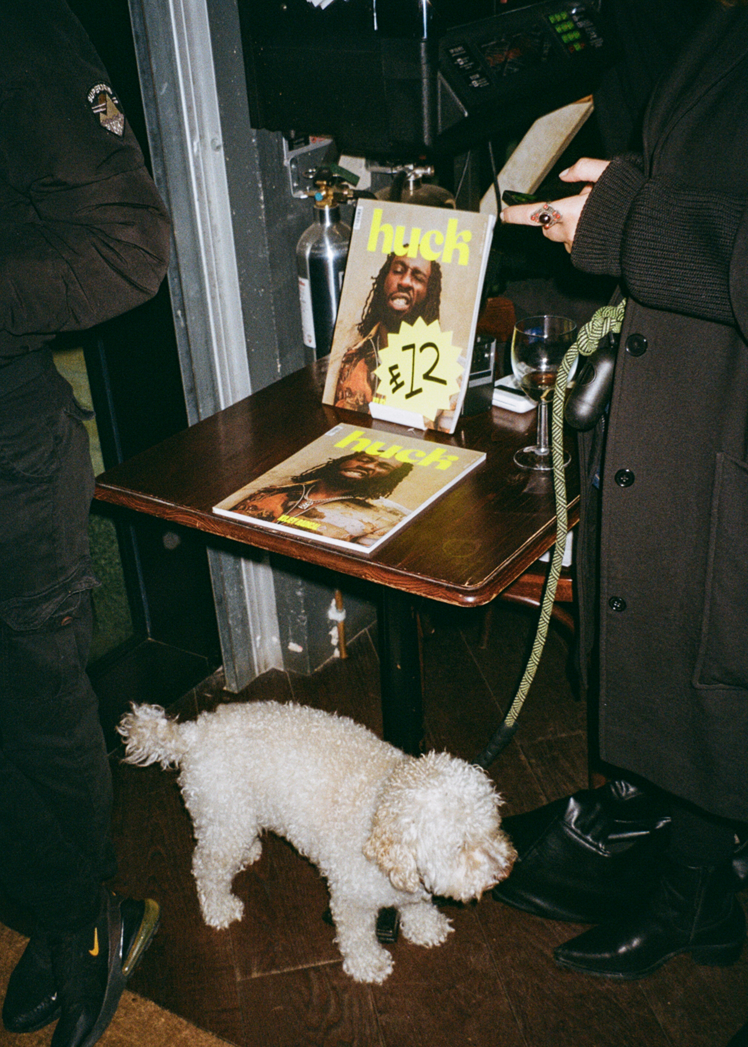 White fluffy dog on wooden floor beneath desk with magazines, glass bottle, and recording equipment in dimly lit room.