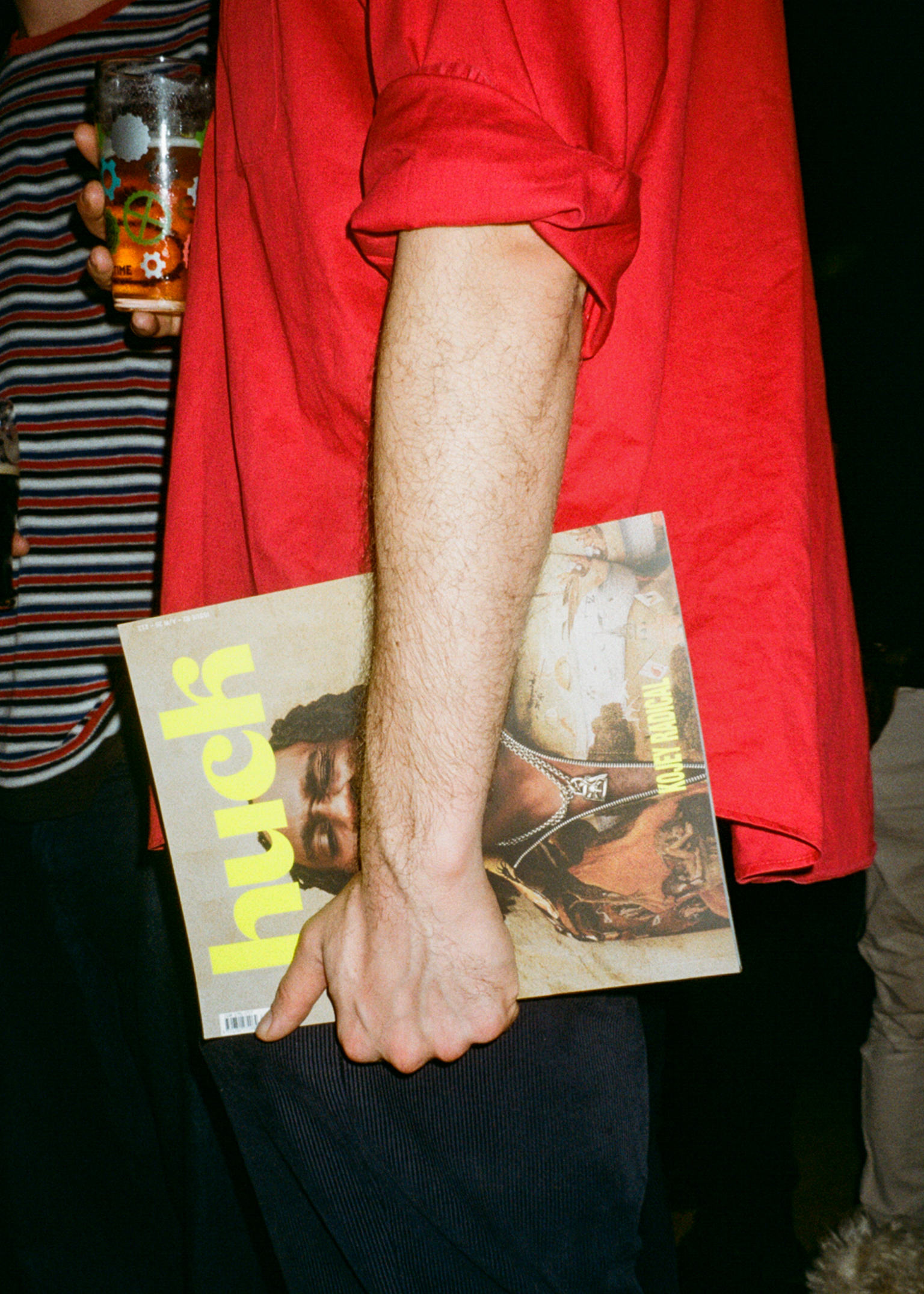Person in red shirt holding magazine and drink, with someone in striped shirt visible in background.