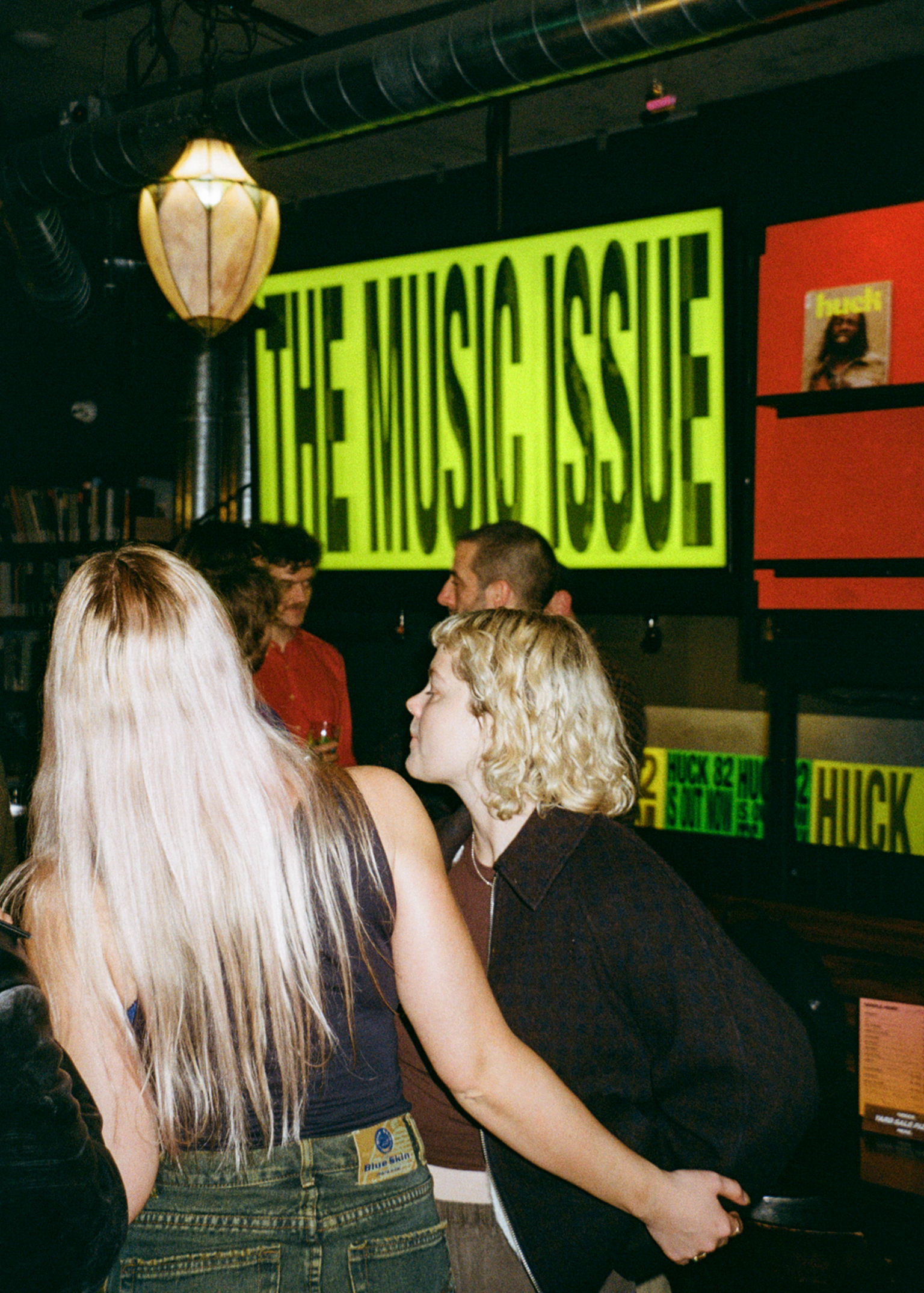 Record shop interior with customers browsing. Large lime green "THE MUSIC ISSUE" sign on wall, orange sections visible. Industrial ceiling with pendant lamp.