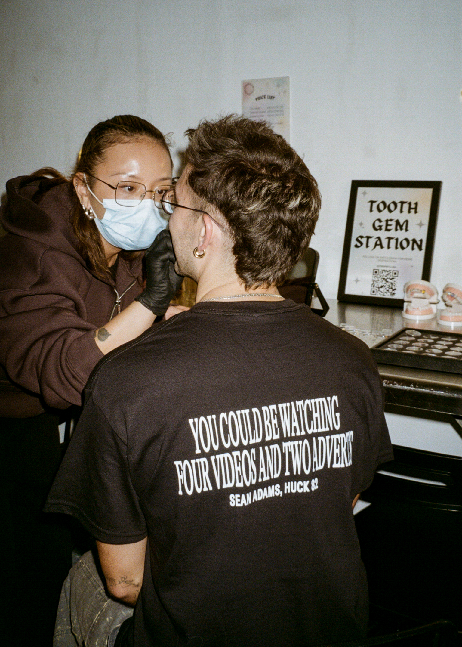 Dental professional in face mask and glasses examining patient wearing black t-shirt reading "YOU COULD BE WATCHING YOUR VIDEOS AND I'M DEAD" at tooth gem station.