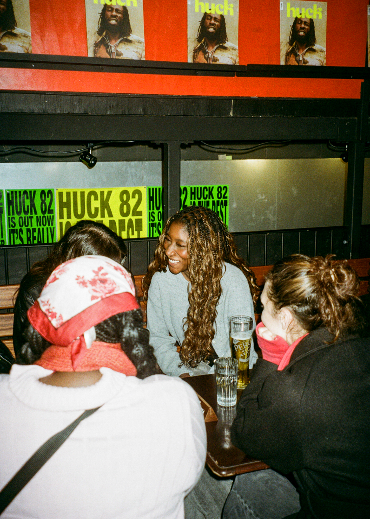 Three people sitting at pub table with drinks. Woman with long dreadlocks in centre wearing grey top. Green "HUCK 82" posters on wall behind.