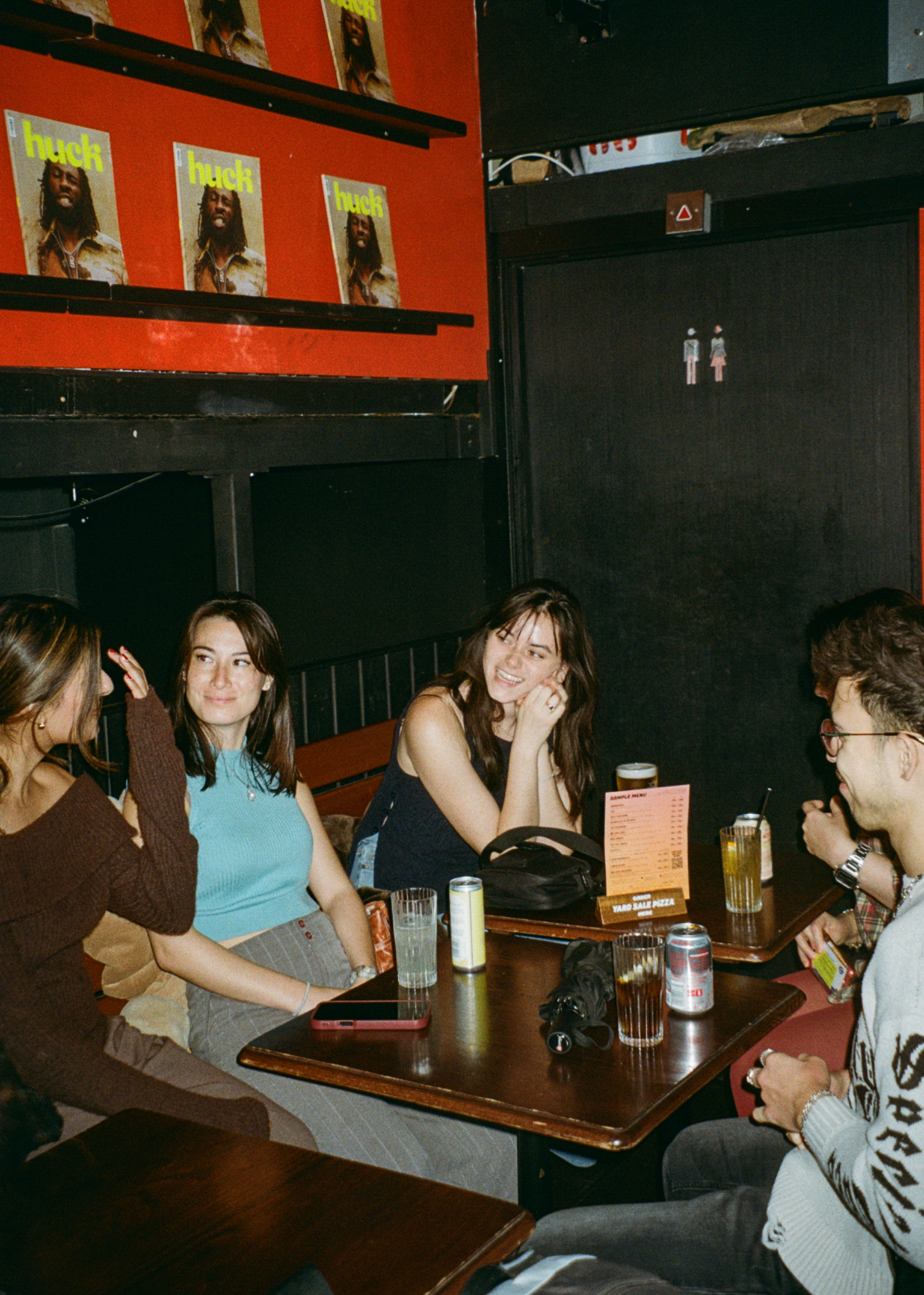 Group of young people sitting at table in pub with drinks, orange walls with posters, dark ceiling beams visible above.