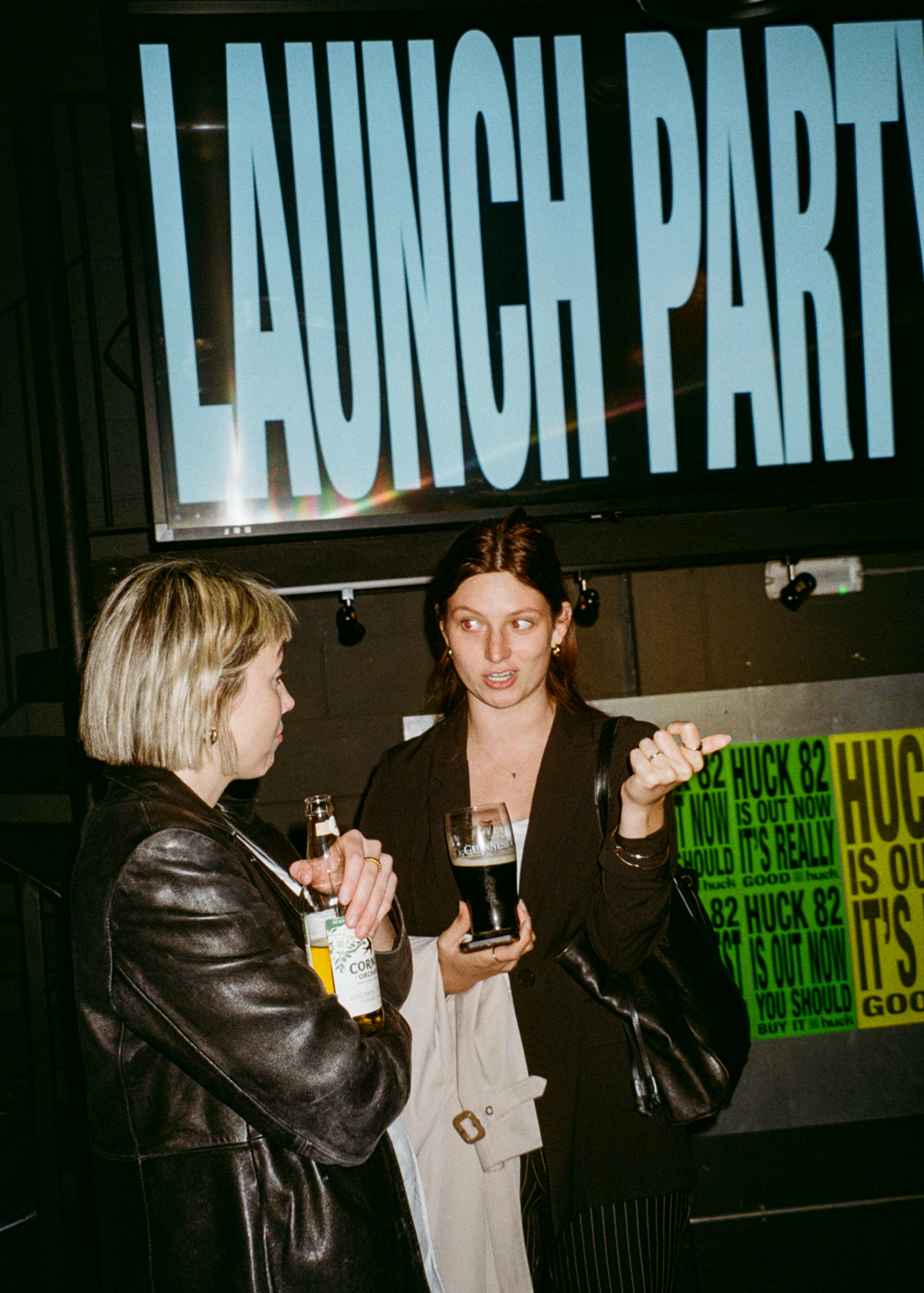 Two women holding drinks beneath large "LAUNCH PARTY" sign, with green promotional posters visible on right side.