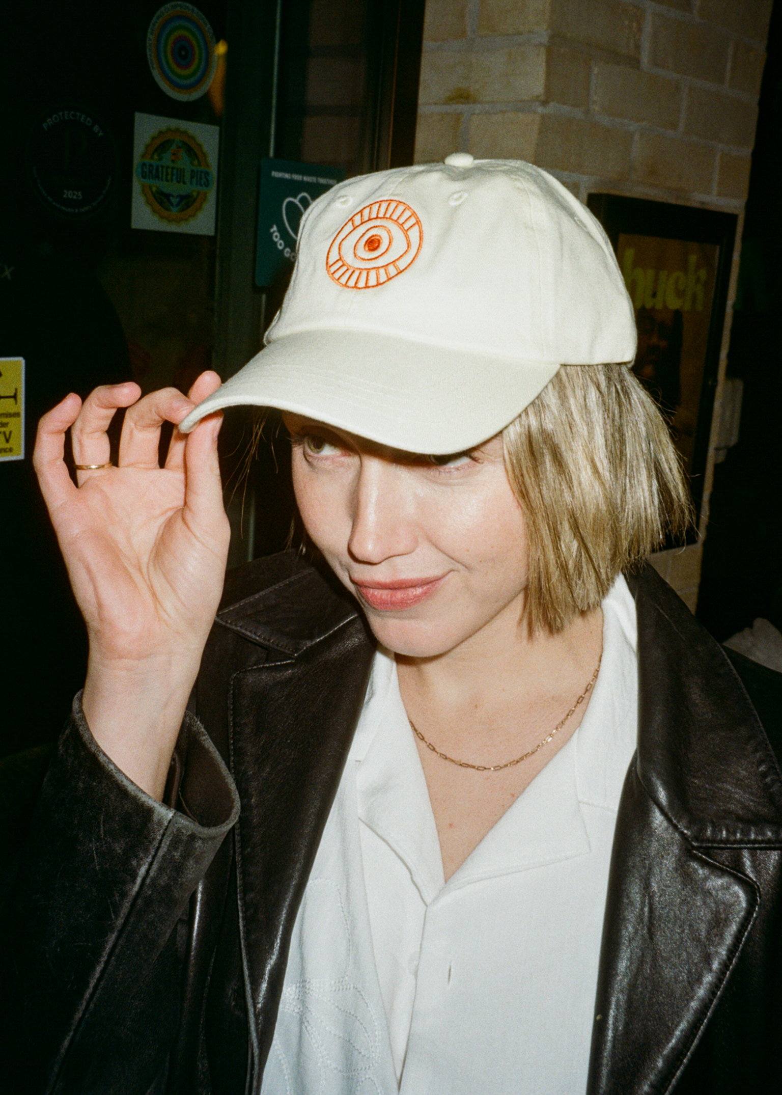 Person in white baseball cap with orange eye logo, touching cap brim, wearing black leather jacket and white shirt with gold necklace.