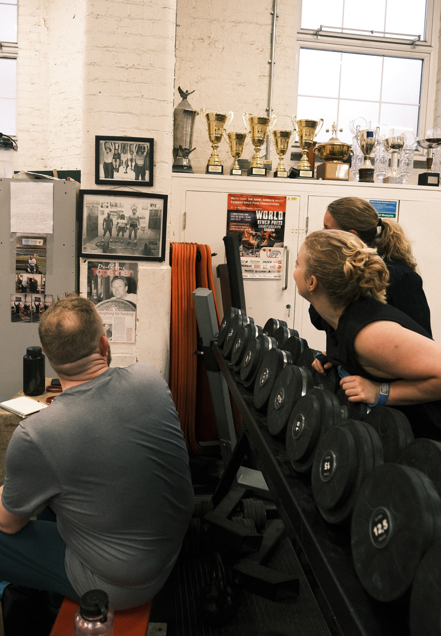 Two people examining black equipment on tripod in room with white brick walls, golden trophies on shelf, and framed photographs.