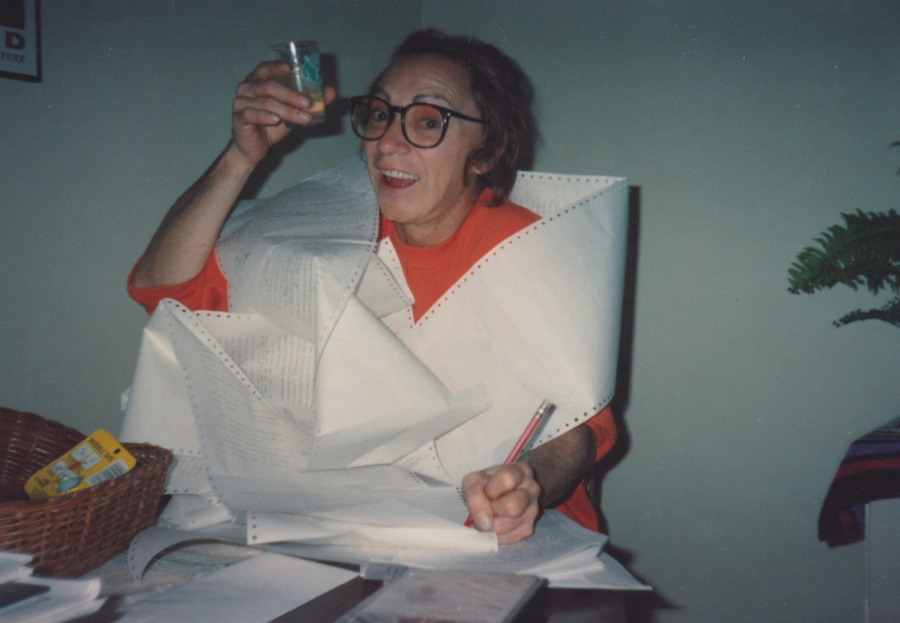 A smiling person wearing glasses and a red jumper holds a mug and sits at a desk with papers.