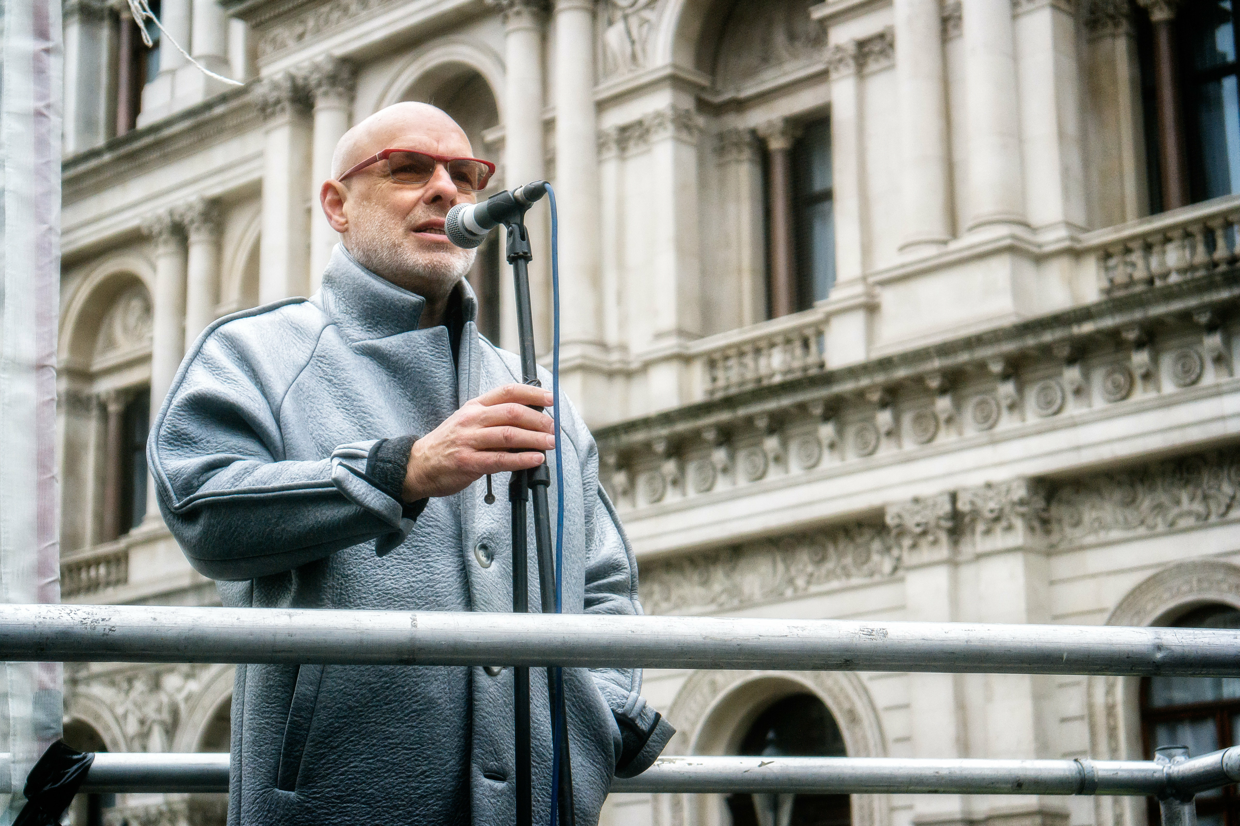 Bald man with sunglasses in grey coat speaking at microphone on outdoor stage, ornate classical building with columns behind
