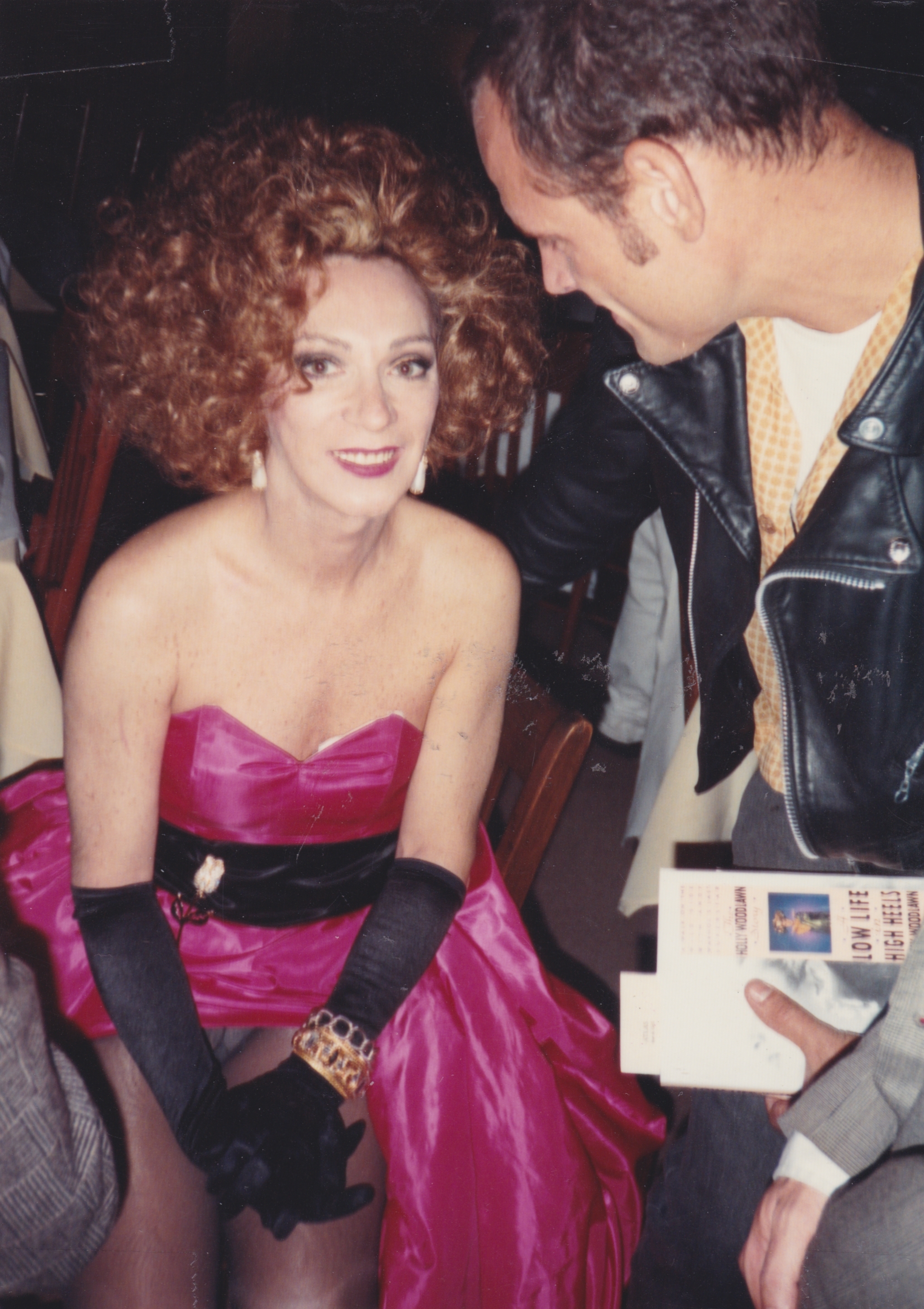 Glamorous woman in a pink satin gown with voluminous curly hair, standing next to a man in a leather jacket.
