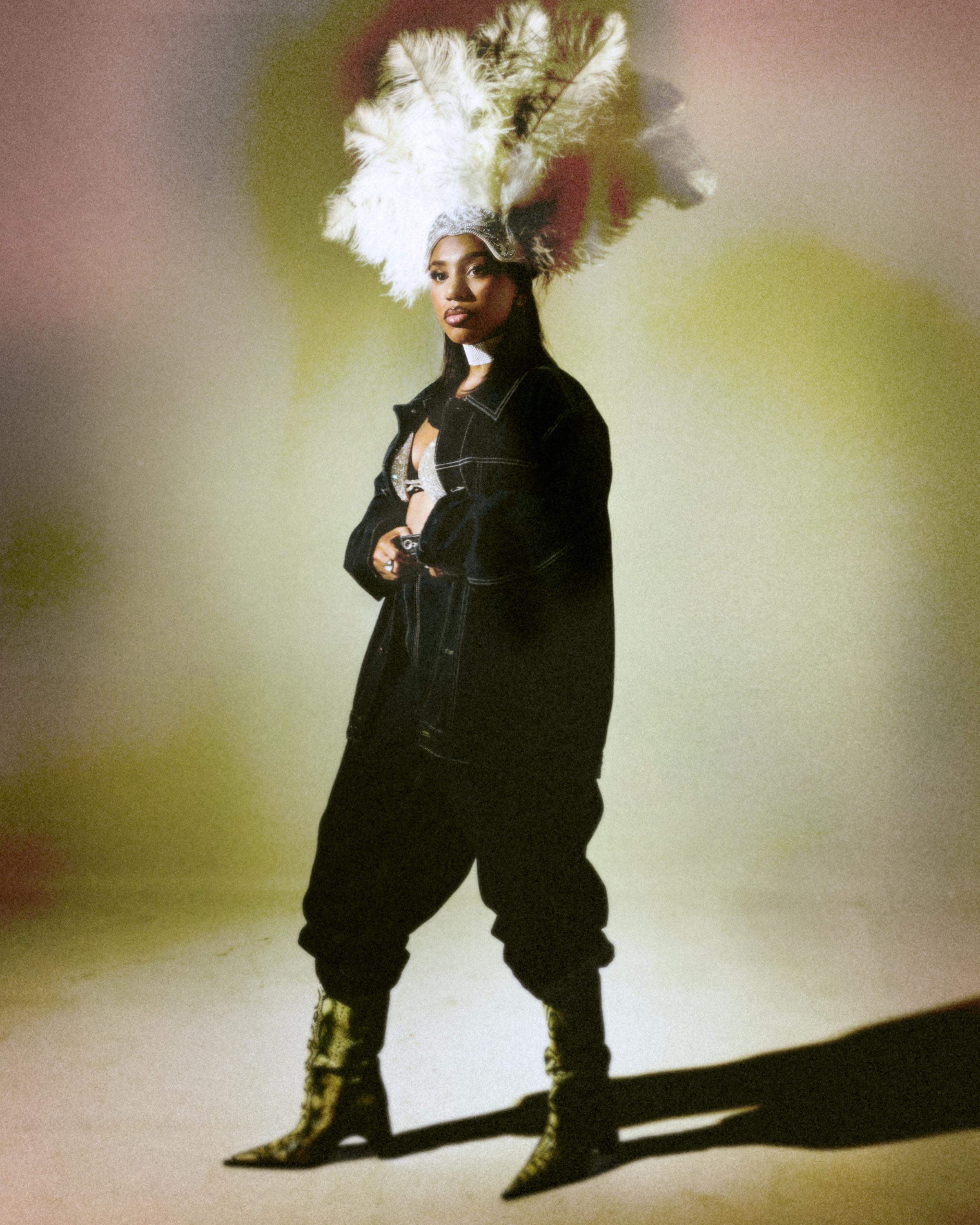Dramatic portrait of person wearing large feathered headdress and dark outfit, standing in colourful lighting.