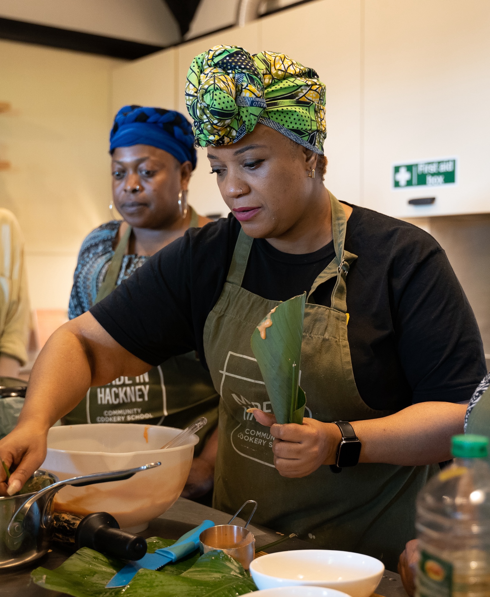 Two women cooking in kitchen wearing green aprons. Woman in foreground has colourful headwrap, woman behind wears blue headwrap.