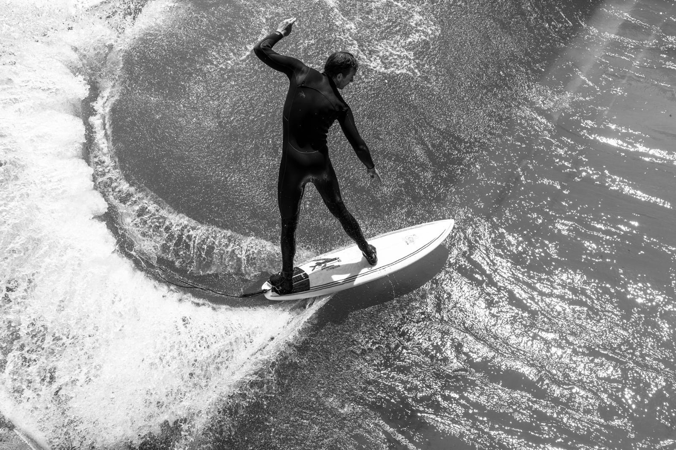 Black and white aerial view of surfer in wetsuit riding wave on surfboard, surrounded by white foam and dark water.