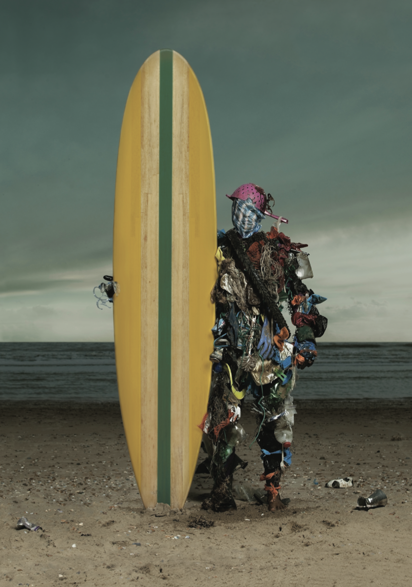 A person in an ornate, colourful costume holding a yellow surfboard on a beach.