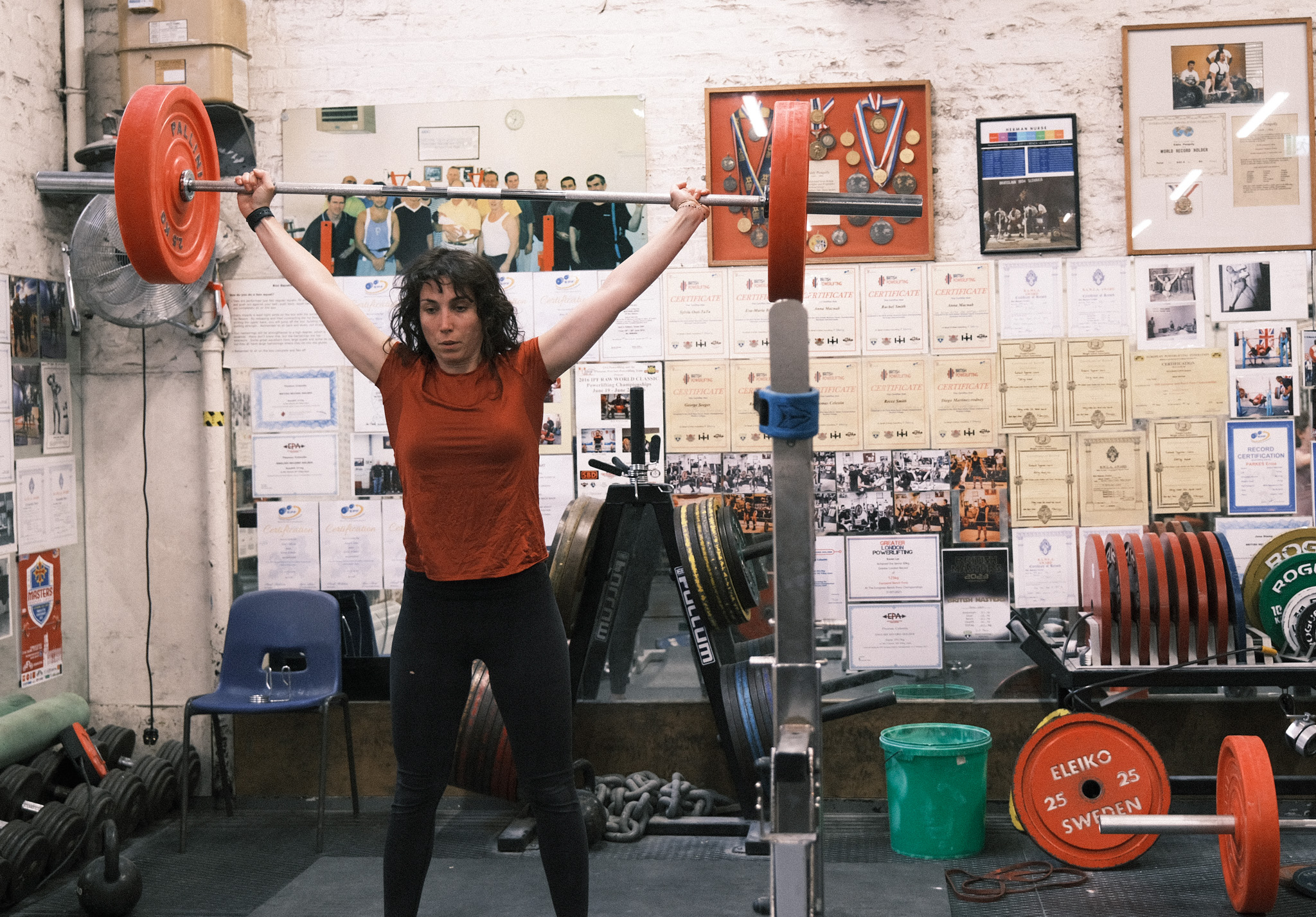 Woman lifting barbell overhead in gym with white walls covered in photographs and documents, orange weights visible.
