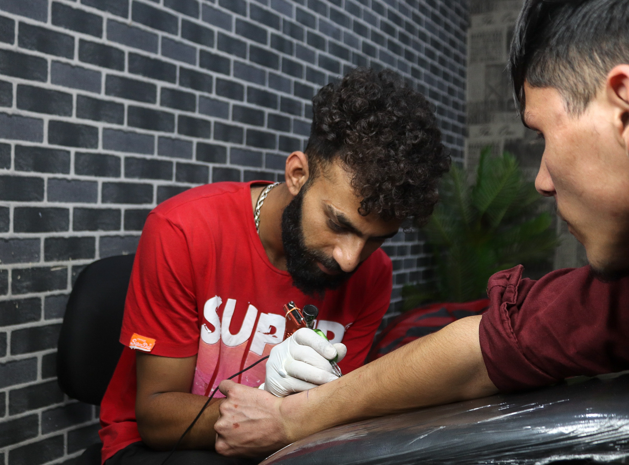 Two young men, one wearing a red "SUPER" t-shirt and the other a maroon shirt, having a conversation against a brick wall.