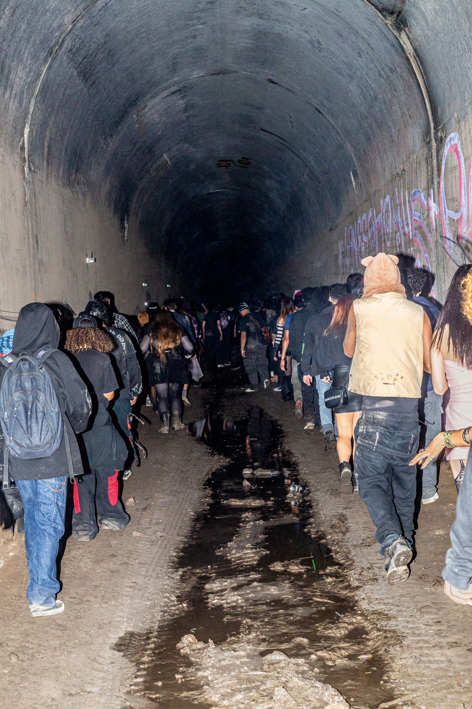 Crowd of people walking through concrete tunnel with graffiti on walls, water puddles on ground, curved ceiling receding into darkness.