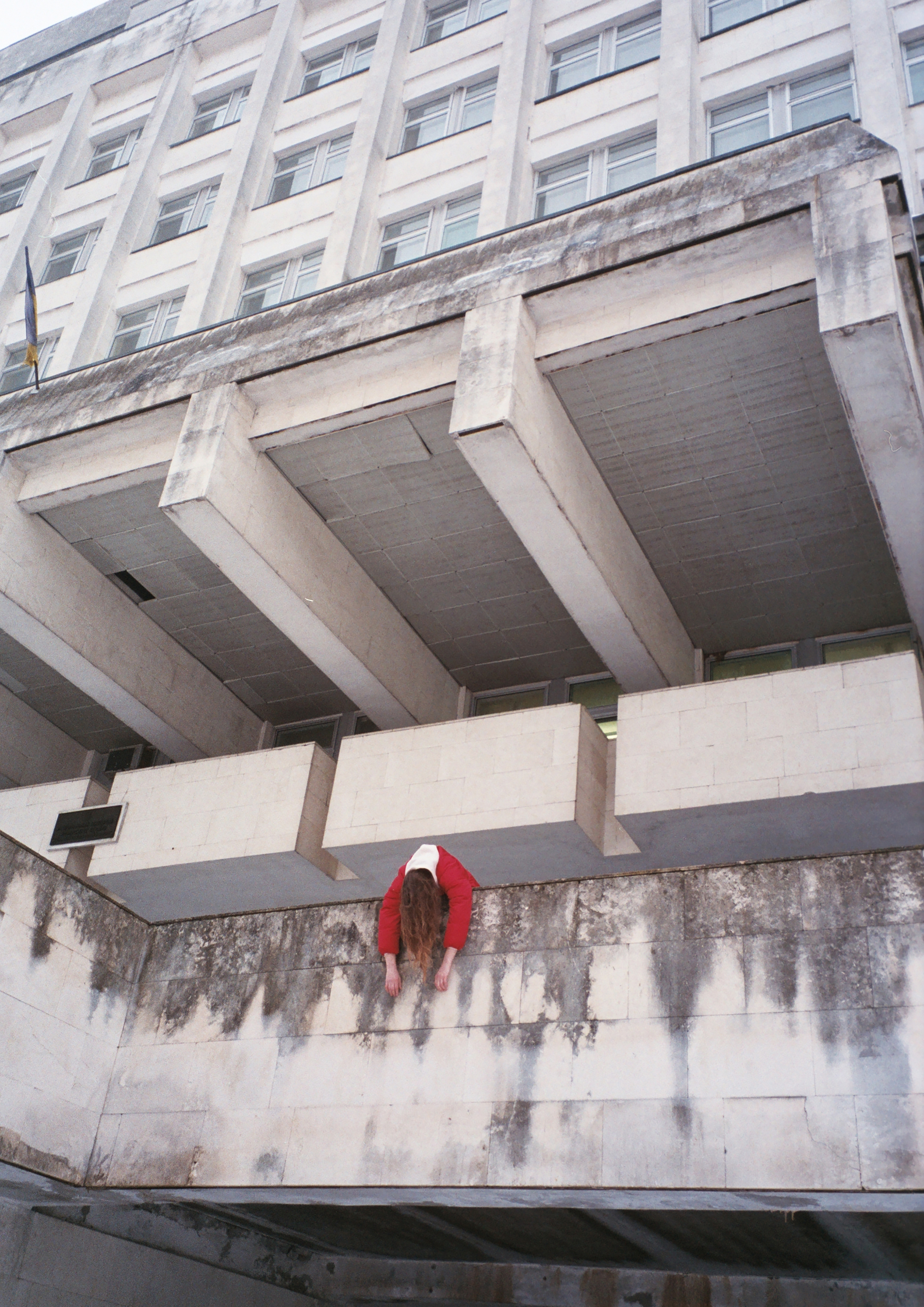 Large grey concrete building with recessed entrance and figure in red clothing dangling from its exterior.