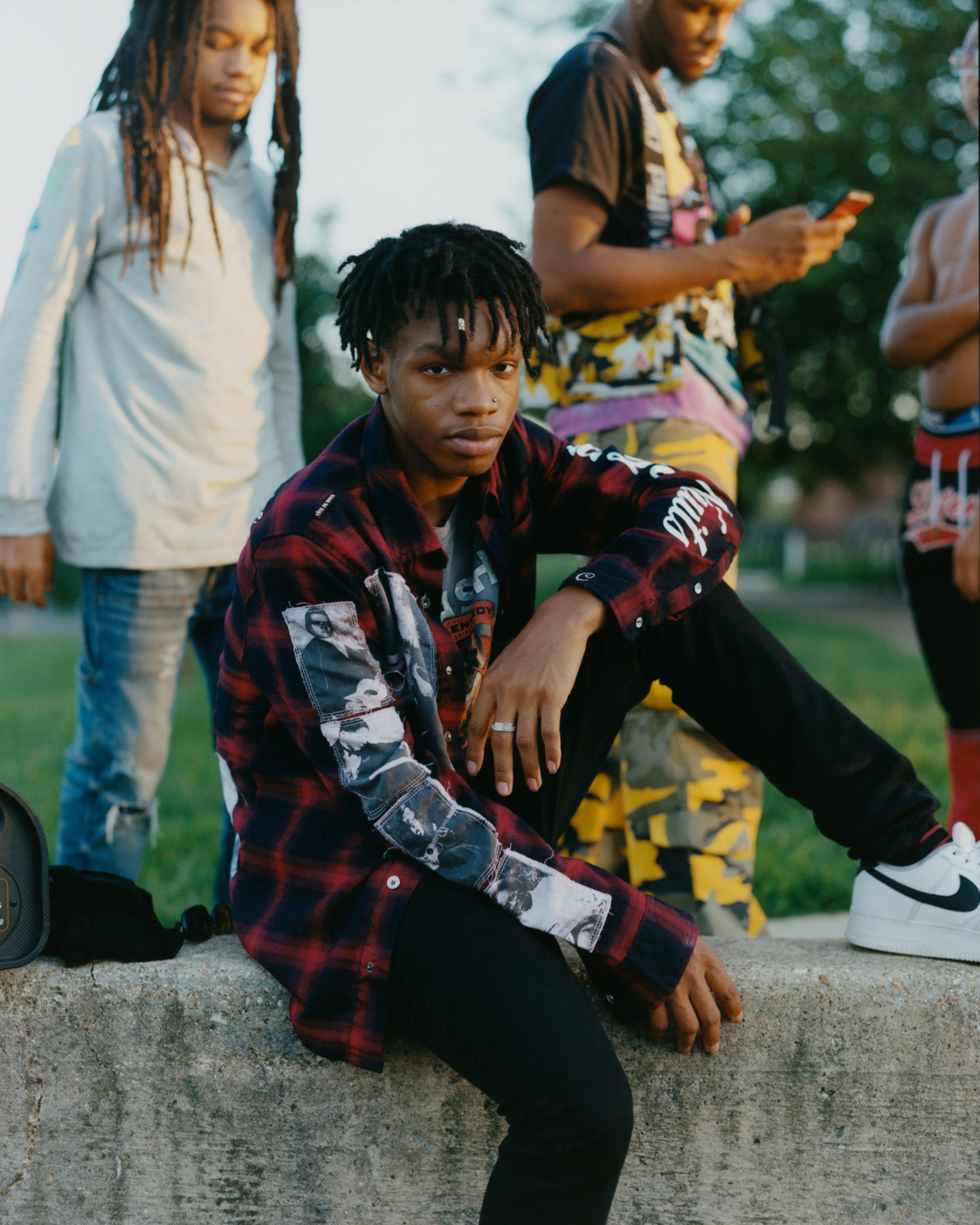 Young man with dreadlocks crouching on pavement wearing red plaid shirt with patches and black trousers, others standing behind.