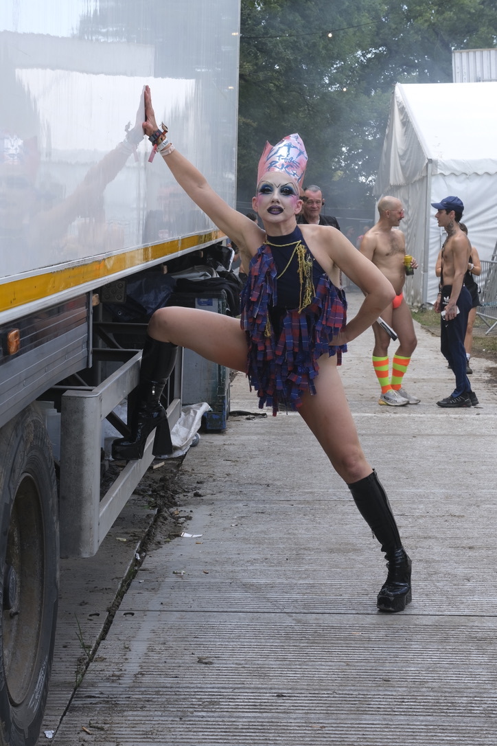 Drag performer in colourful headpiece and fringed outfit poses dramatically with one leg raised beside lorry at outdoor event.