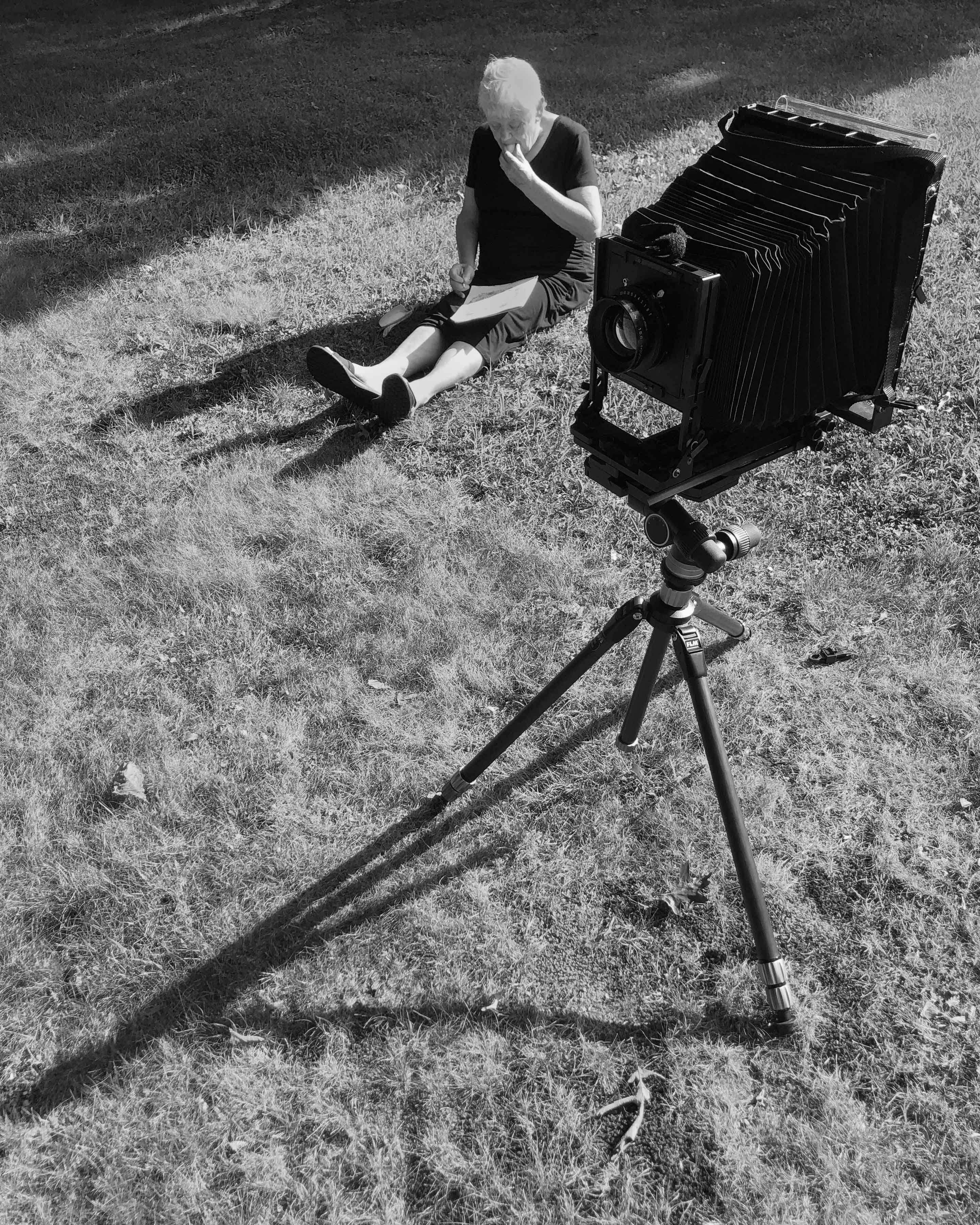 Black and white image showing large format camera on tripod in foreground, woman sitting on grass in background reading book.