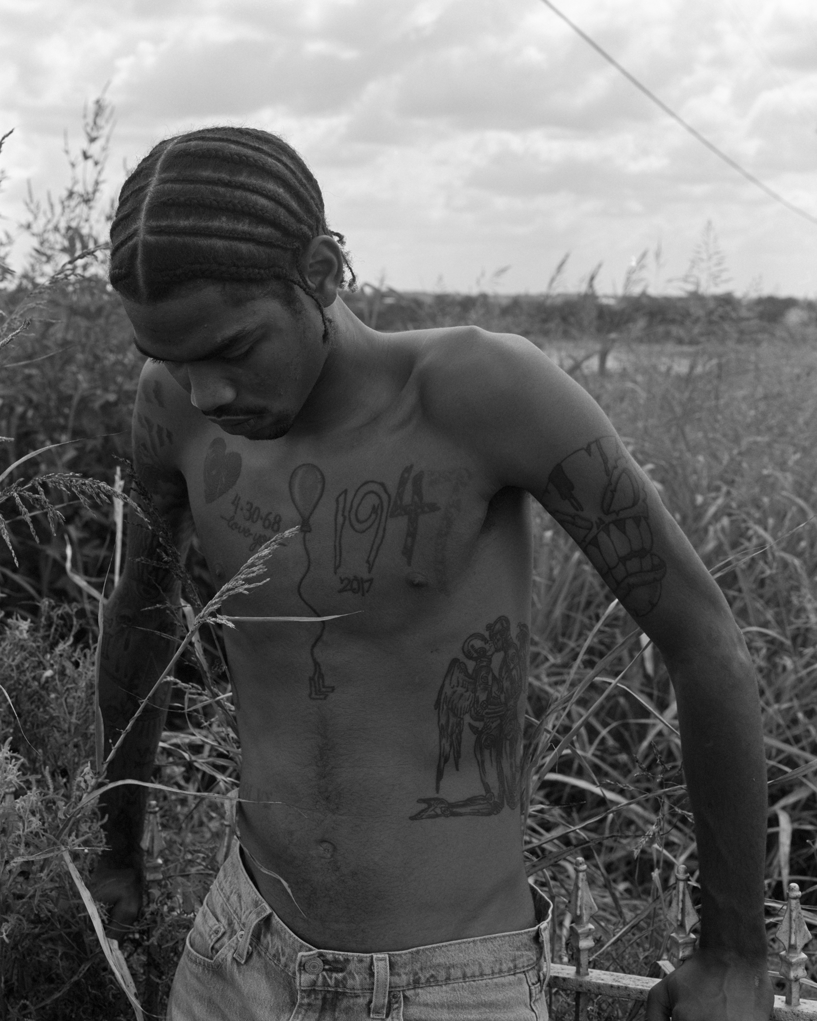 Black and white photograph of a shirtless man with distinctive braided hairstyle, standing amidst tall grass, with text visible on his back.