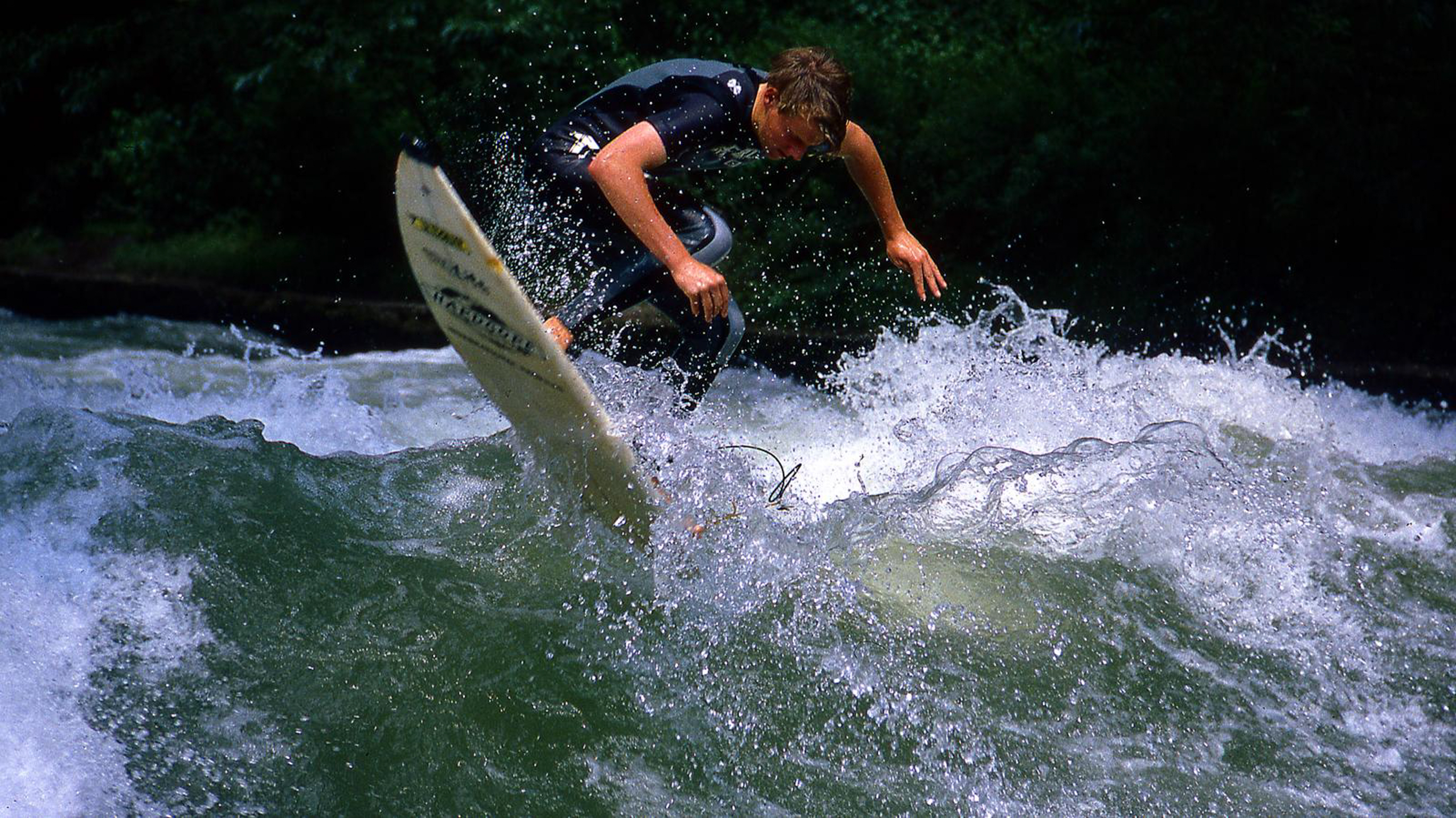 Person in dark wetsuit surfing on white foamy river wave, surrounded by splashing water and green trees in background.