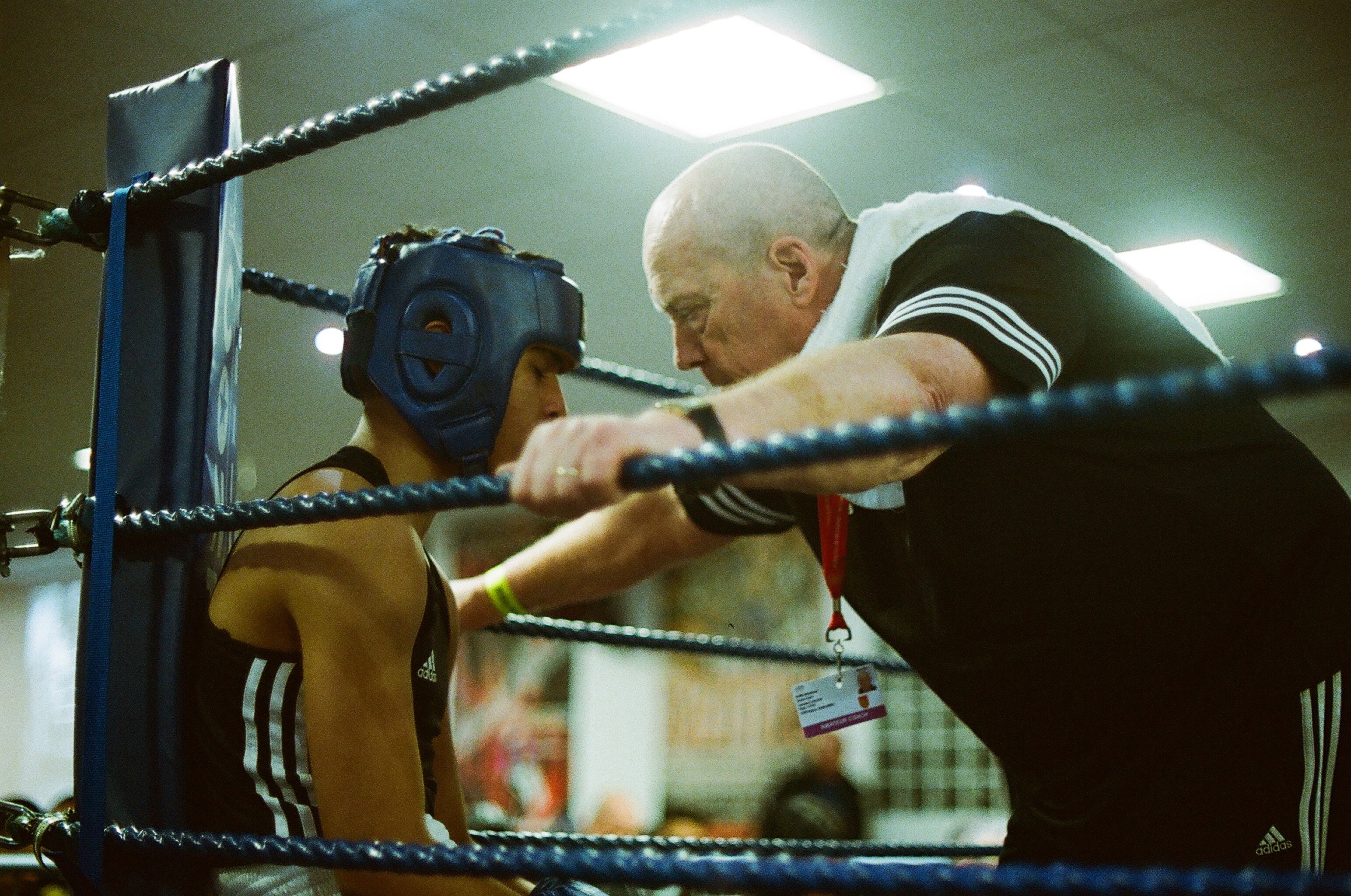Boxing trainer in black tracksuit instructing young boxer wearing headgear in gym ring under bright lights.