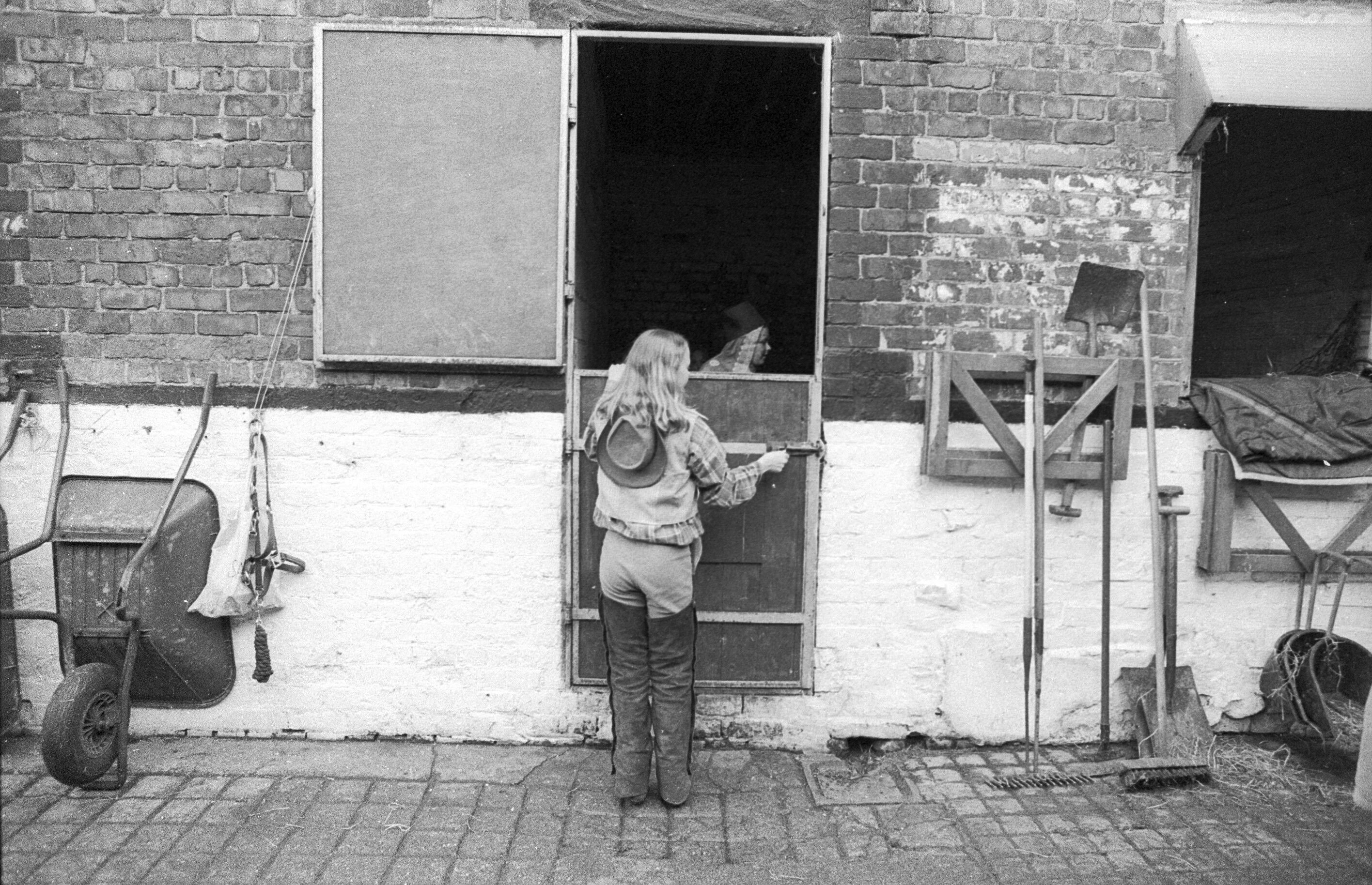Black and white image of woman standing in doorway of dilapidated brick building, holding a mug. Tools and debris on the ground around her.