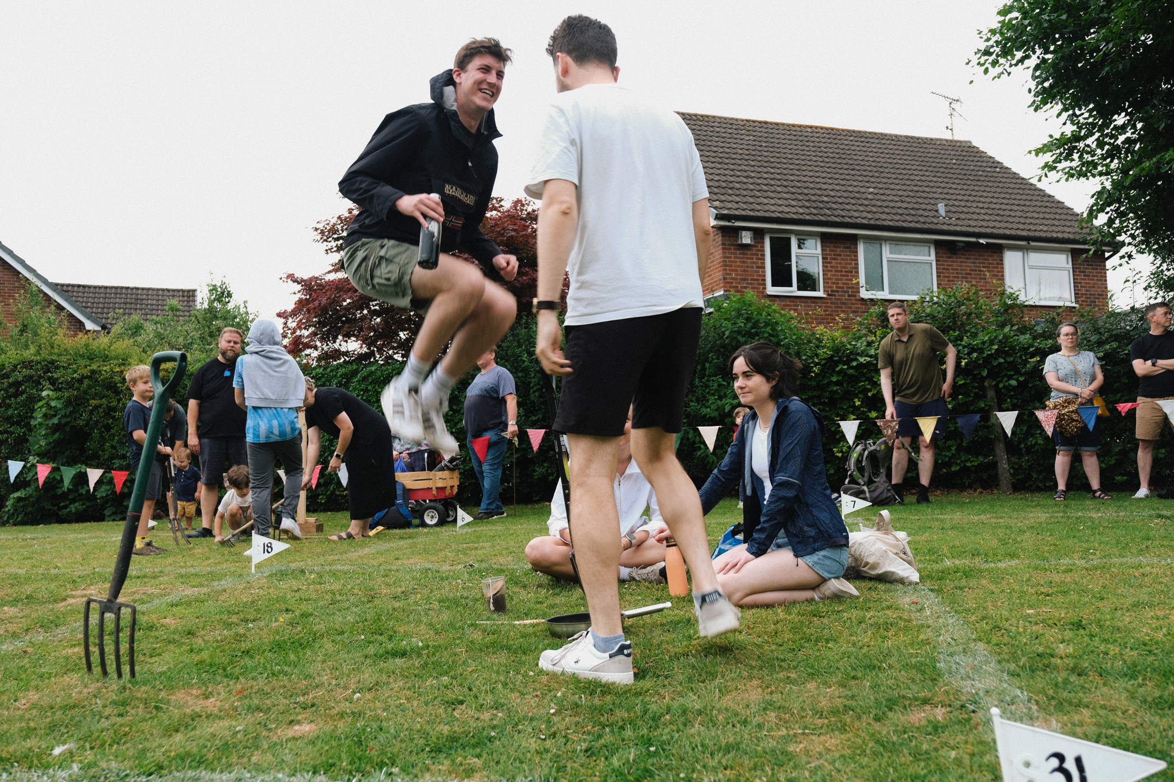 Garden sports day with participants playing games on grass lawn in front of brick houses with spectators watching.