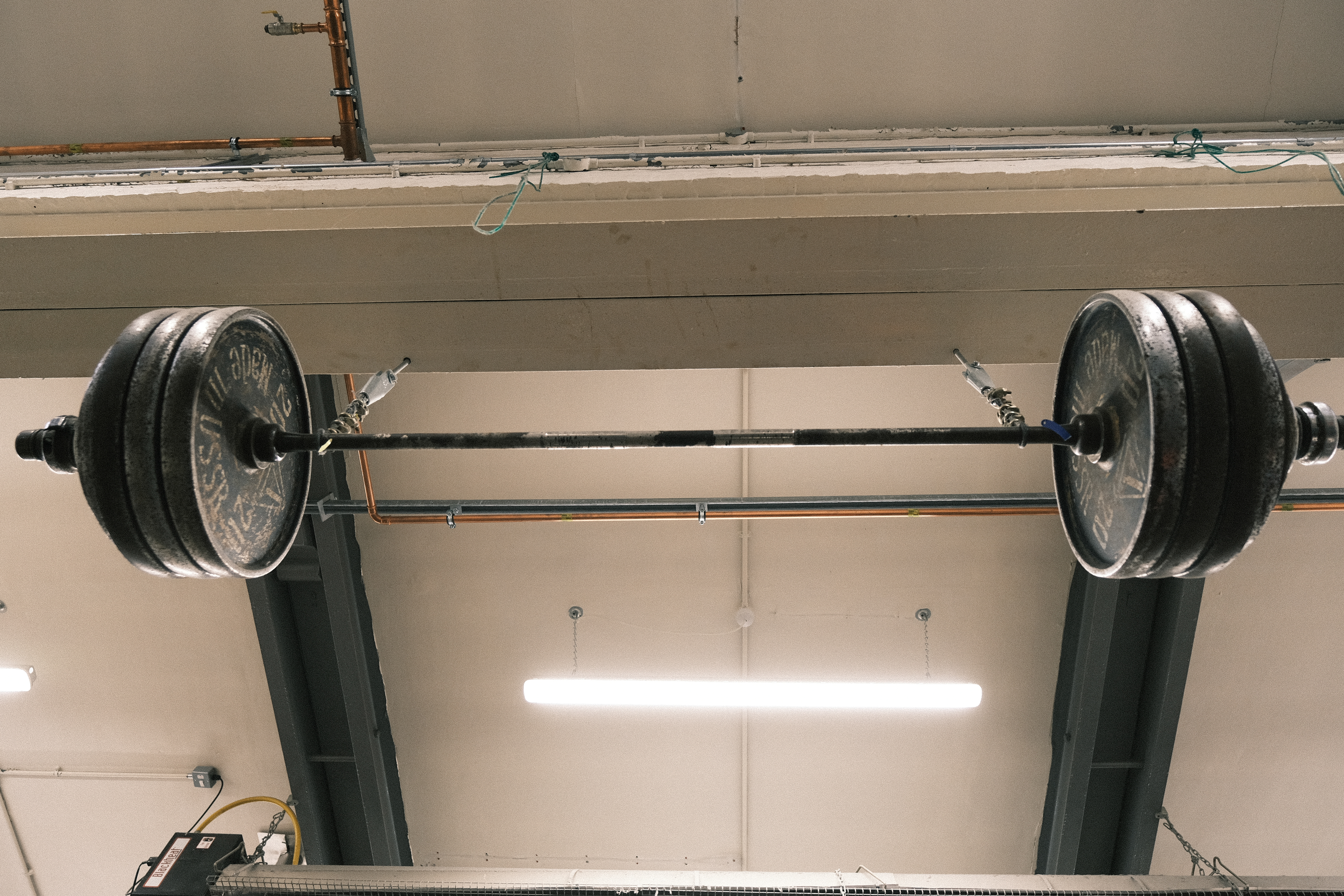 Barbell with weight plates mounted on black squat rack stands in tiled gym room with fluorescent lighting overhead.