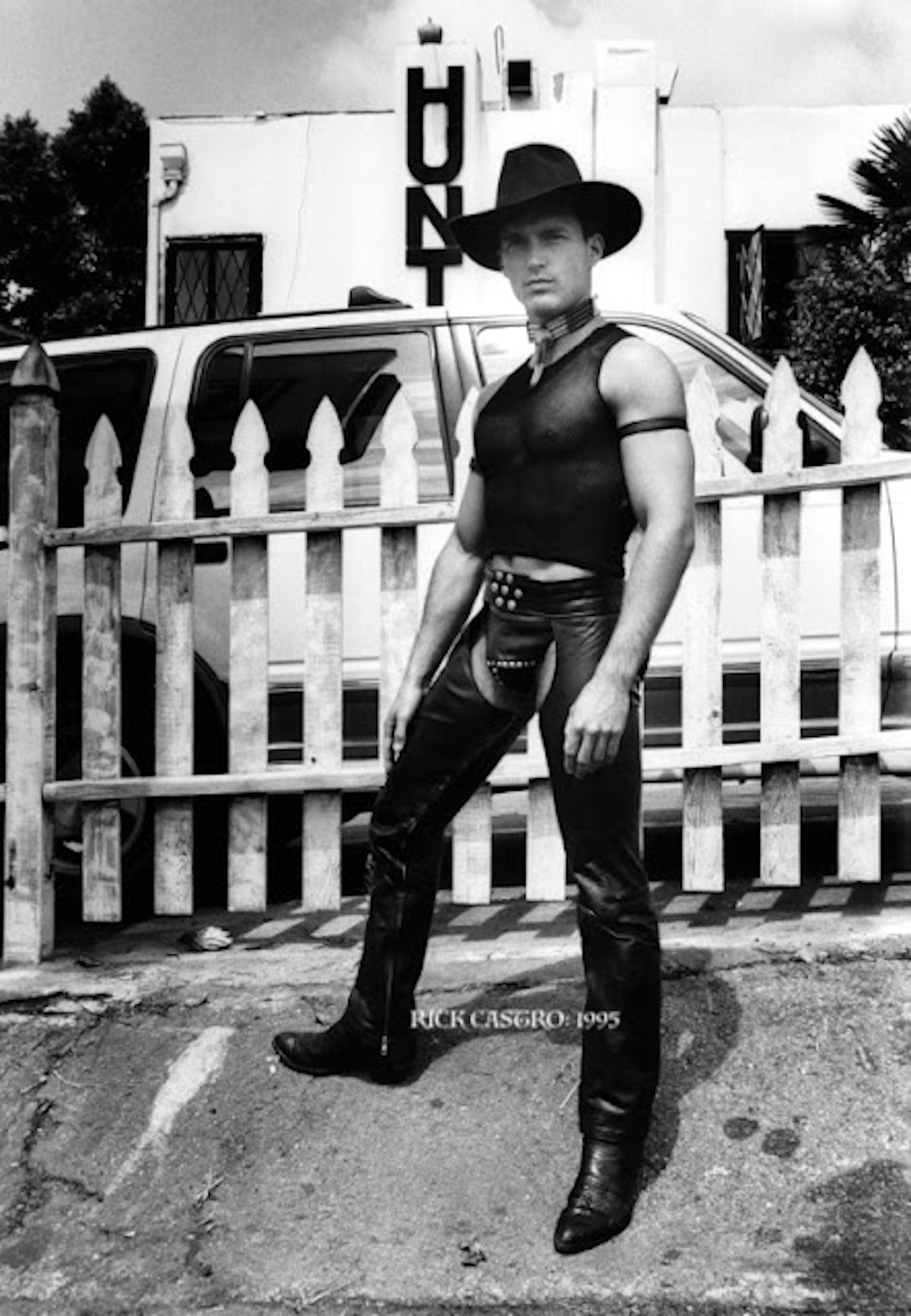 Man in cowboy hat, leather vest and chaps stands beside white picket fence. Black and white image with motel sign visible in background.