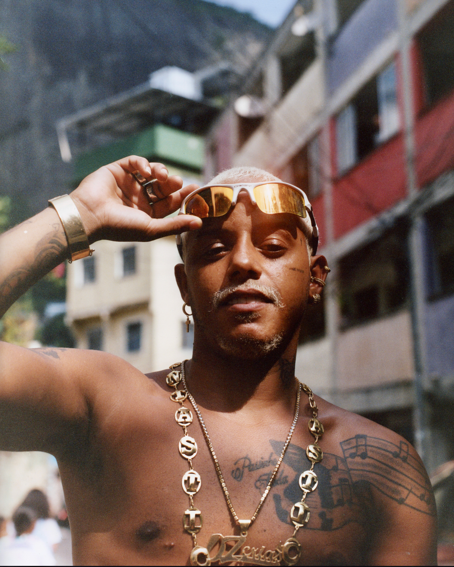 Shirtless man with gold chains and sunglasses shielding eyes from sun, colourful buildings blurred in background.