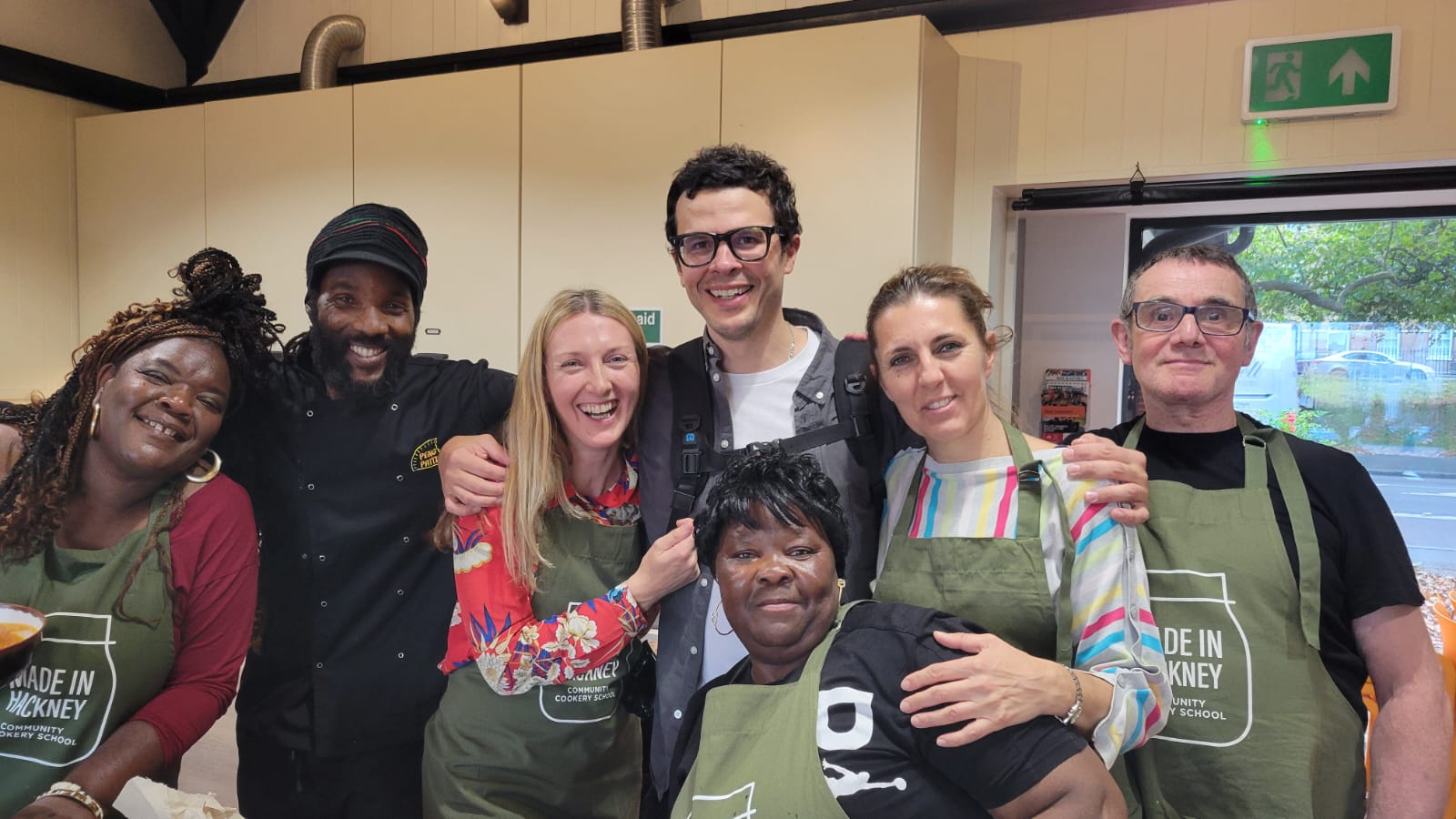 Seven people wearing green aprons pose together in a kitchen, smiling at camera. Emergency exit sign visible on wall behind them.